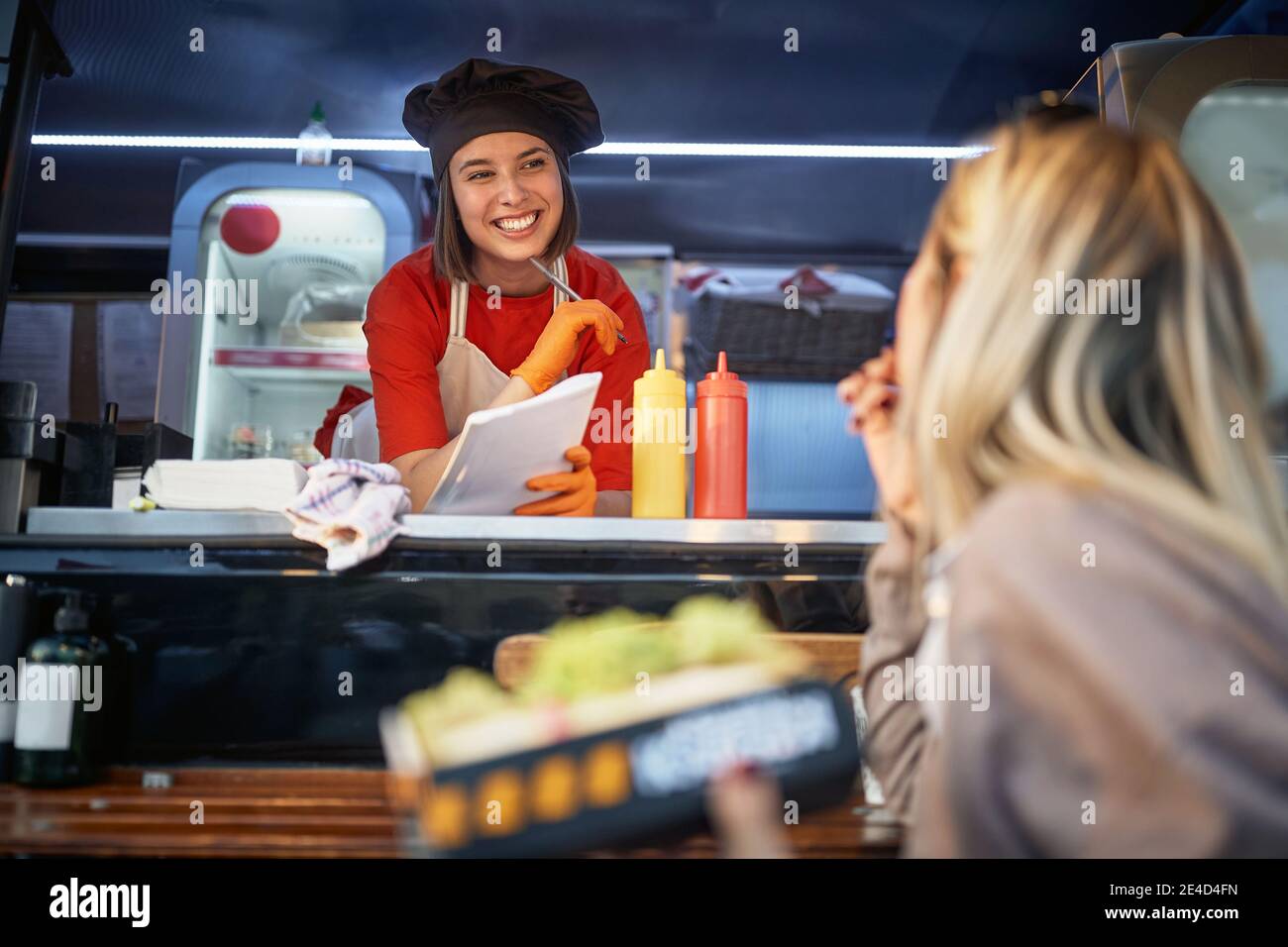 young female listening suggestions from a customer who is eating a sandwich in front of a fast food service Stock Photo