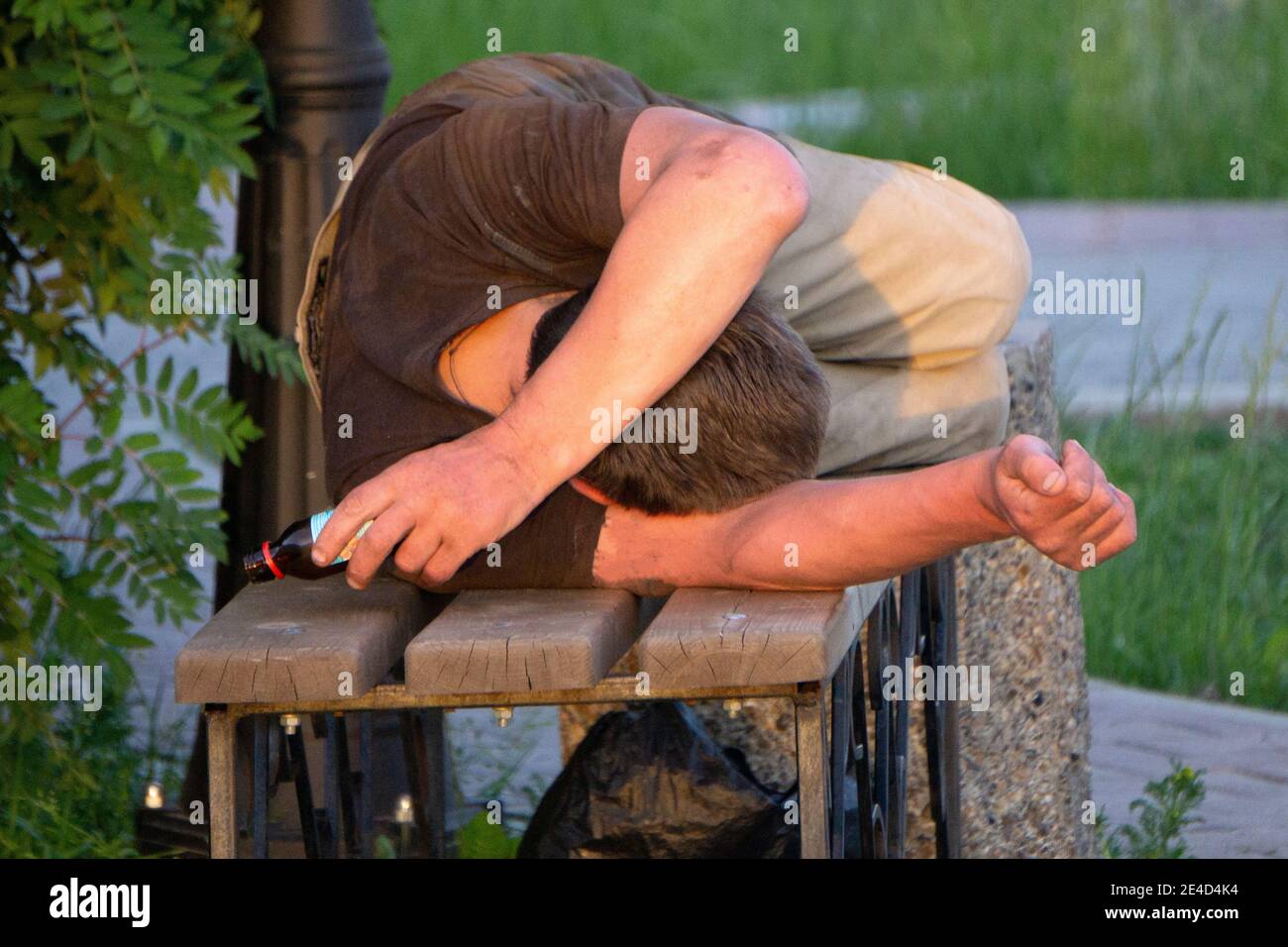 Homeless man fallen down on the ground next to a bench in the park, experiencing lethargy after excessive drinking Stock Photo