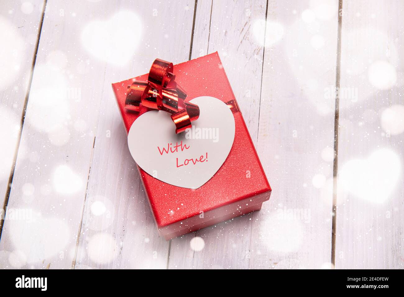 Red box with a big heart-shaped card for a special Valentine gift. White wooden background and bokeh effect all around. Stock Photo
