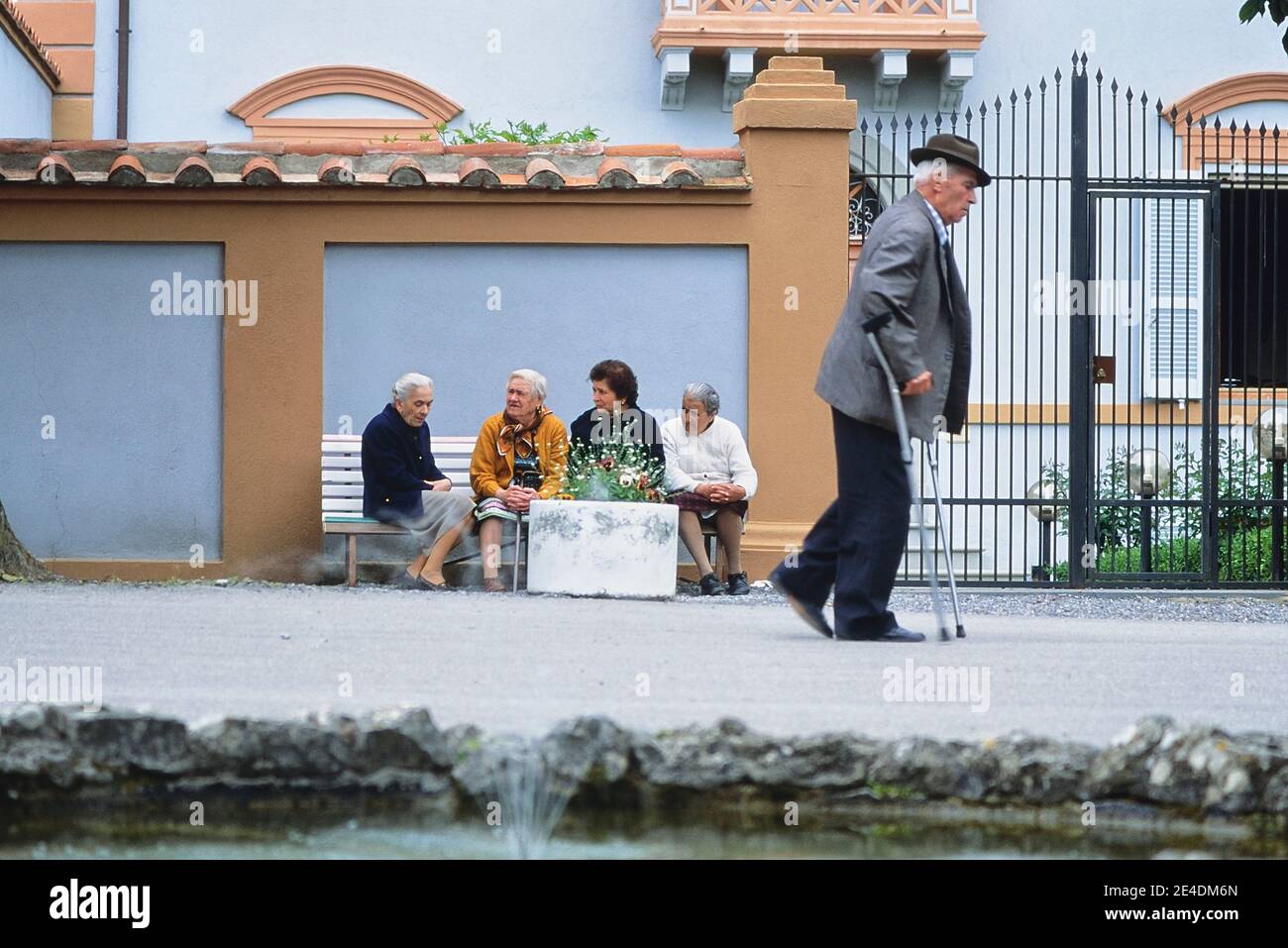 An elderly man with crutches passing four elderly Italian ladies sitting on a public bench having a discussion. Italy Stock Photo
