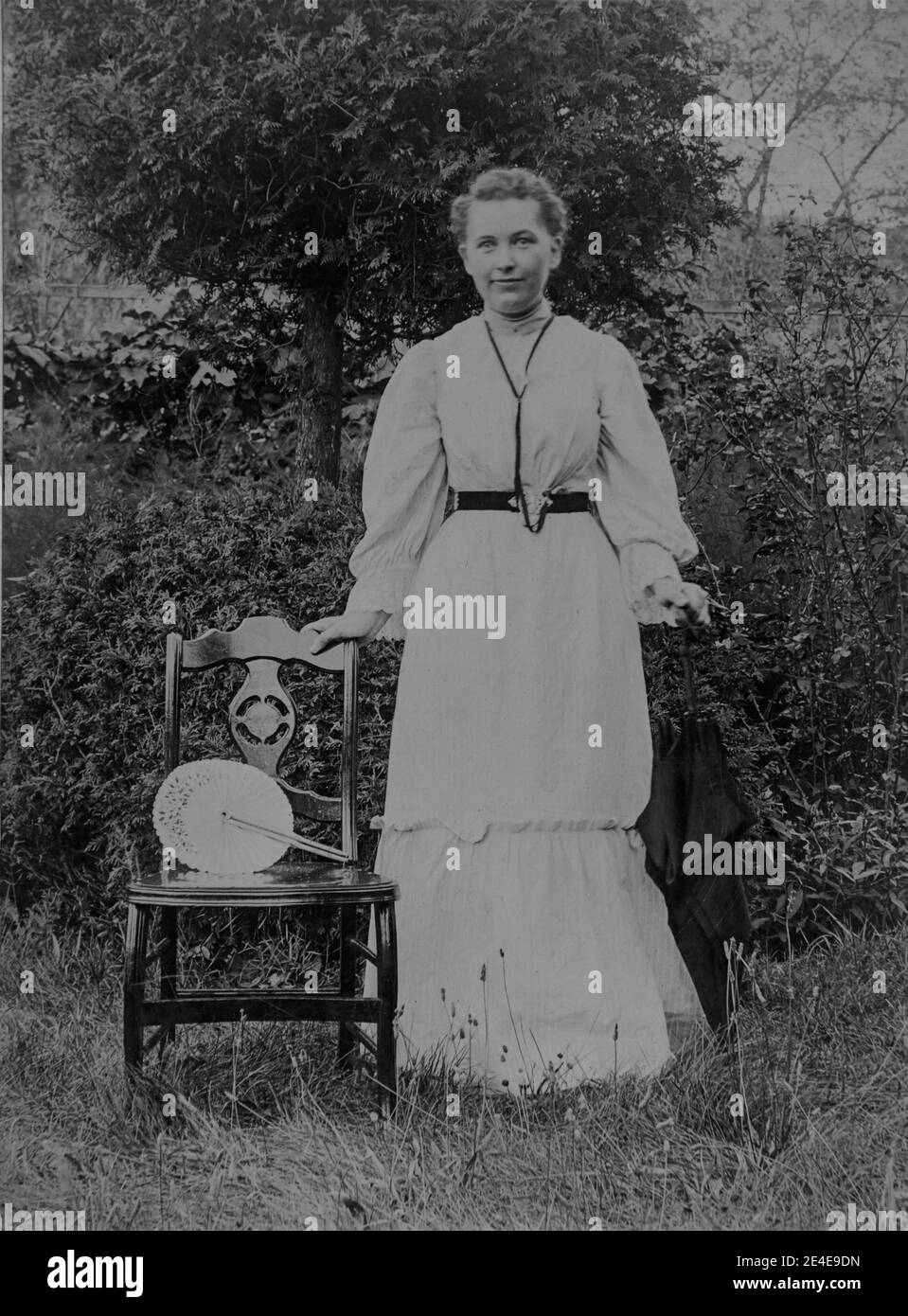 American archive monochrome portrait of a young woman or girl standing next to a chair in a garden with a fan on the chair. The girl is wearing a long white dress and is holding an umbrella. Taken in the late 19th century in Port Byron, NY, USA Stock Photo