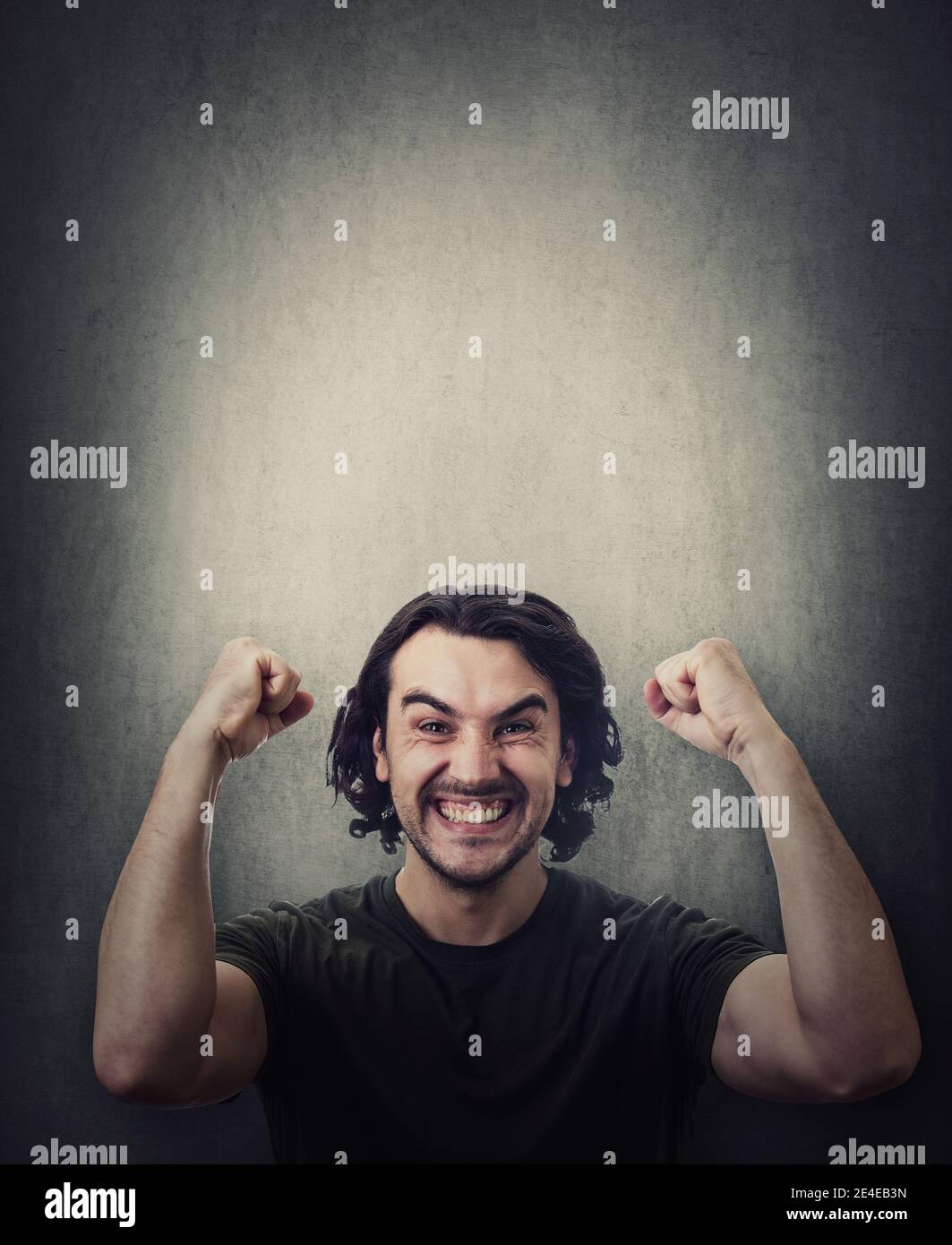 Pleased and positive man, long curly hair style, celebrates success hysterical and passionate. Guy keeps fists tight, raise hands up clenching teeth, Stock Photo