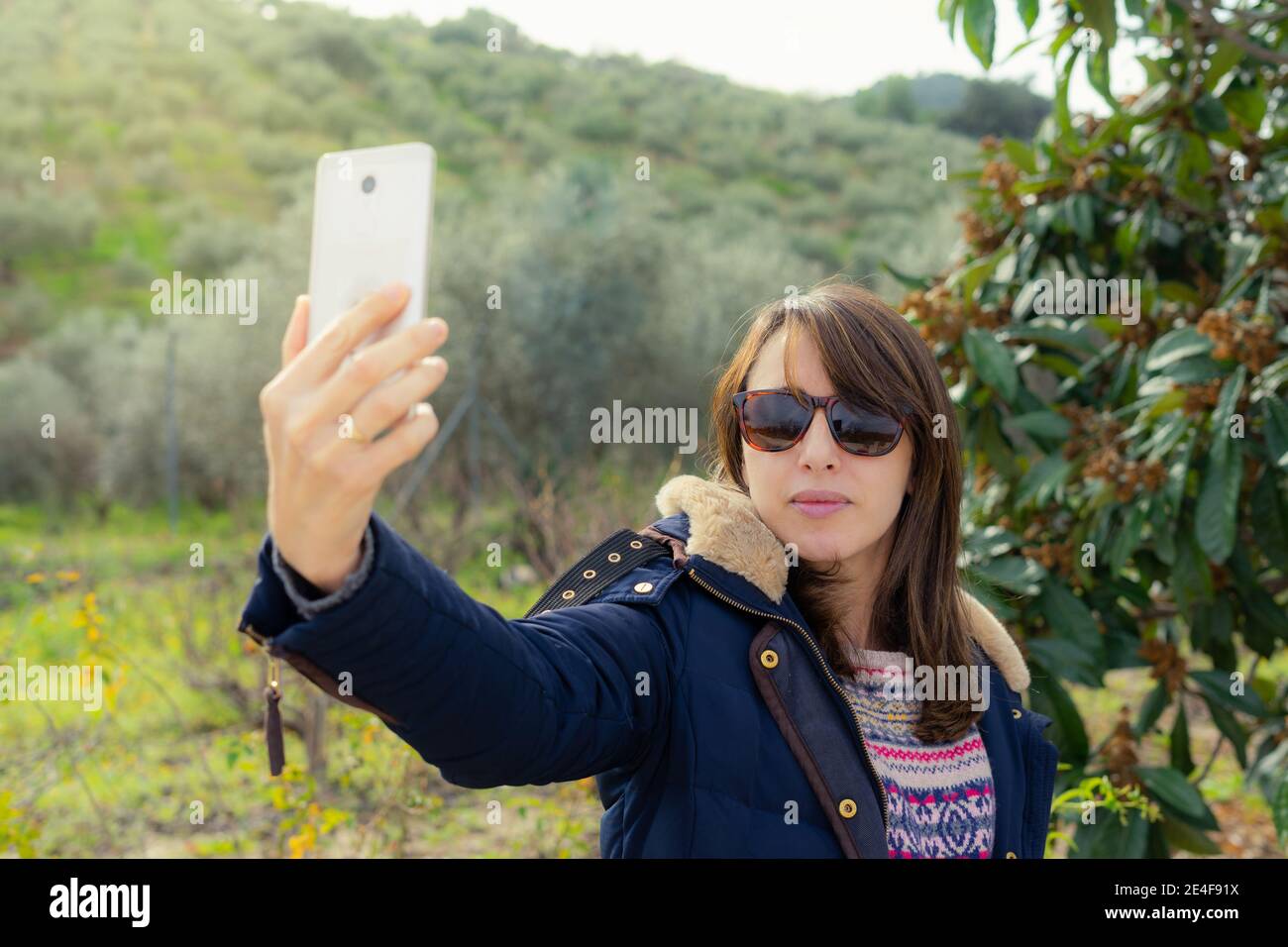 woman takes a selfie in the countryside, wearing a coat and sunglasses. She is smiling and happy. Stock Photo