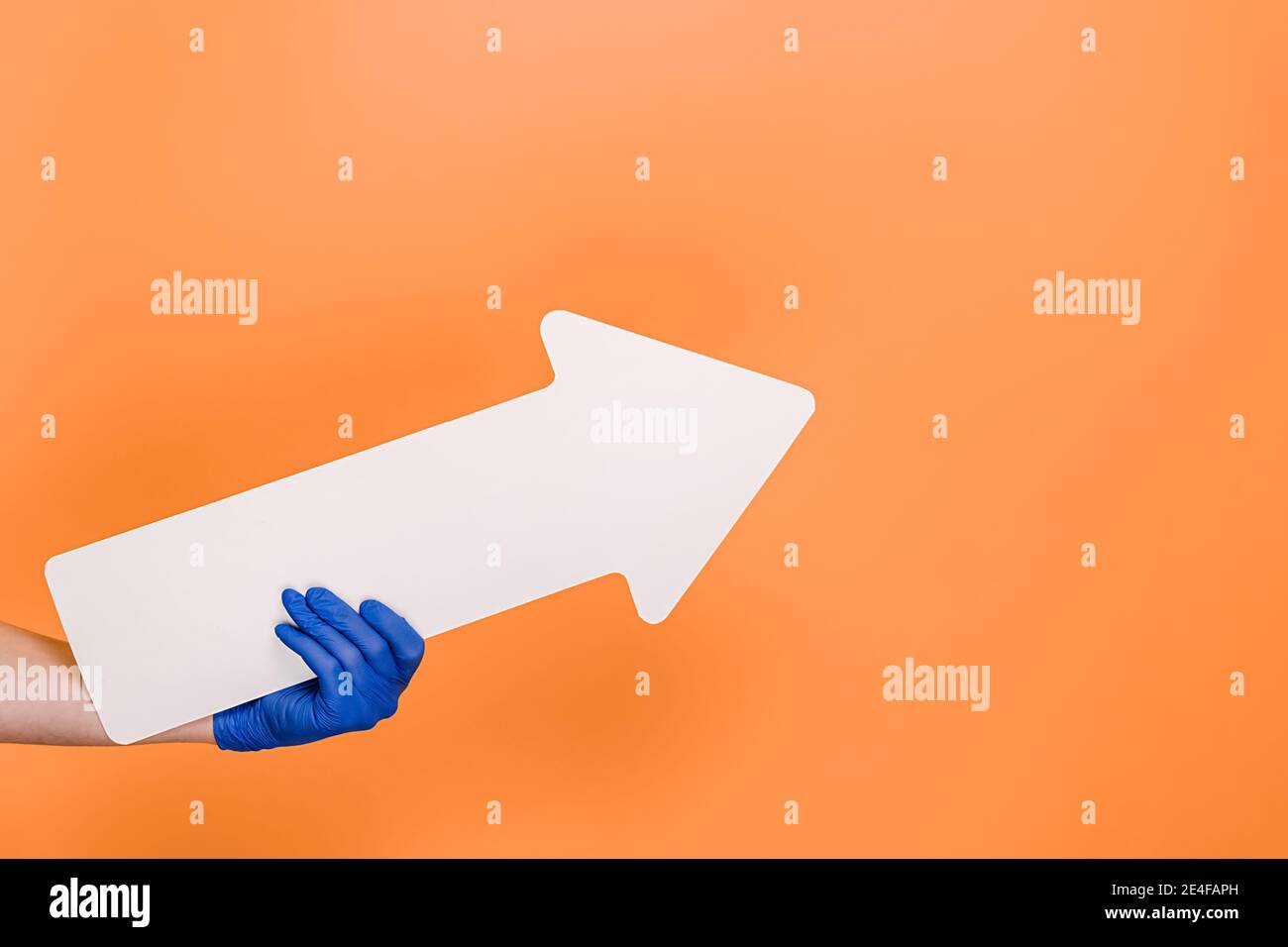 Close up of female in medical protective blue gloves hold white arrow pointing at copy space for advertisement, isolated on orange studio background. Stock Photo