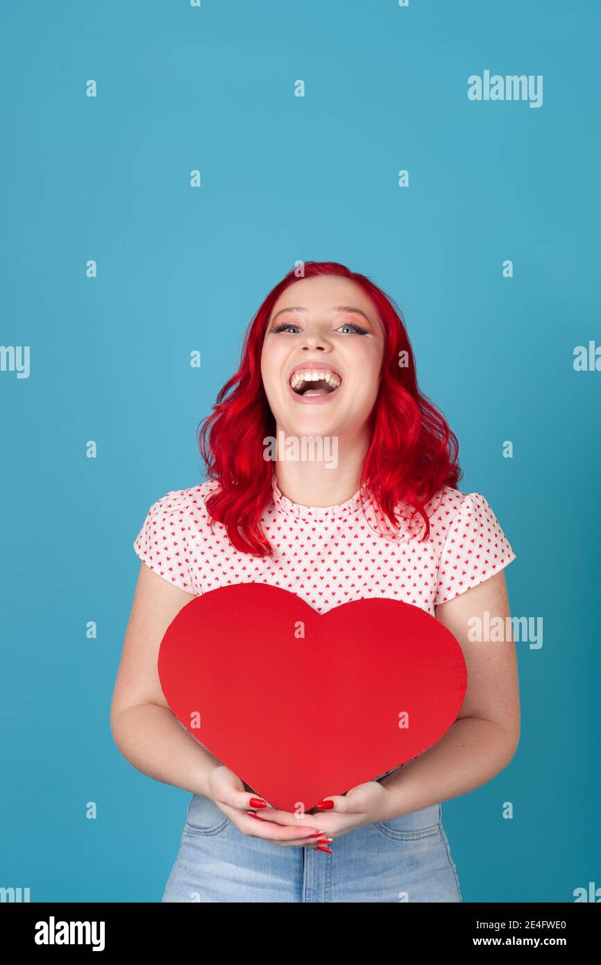 laughing, elated, overjoyed young woman with red hair holds a red paper heart in two hands , isolated on a blue background Stock Photo