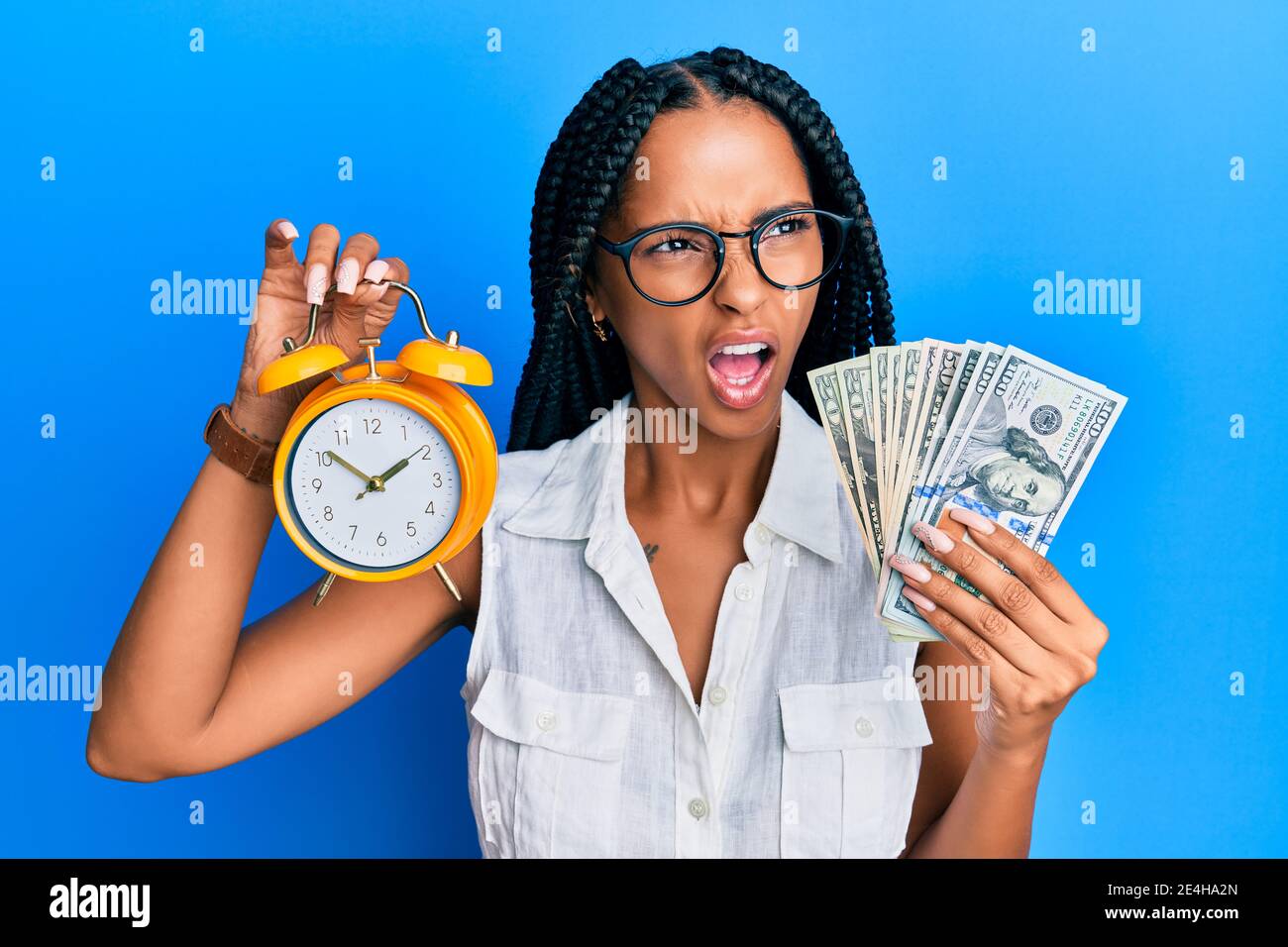 Beautiful hispanic woman holding alarm clock and dollars angry and mad ...