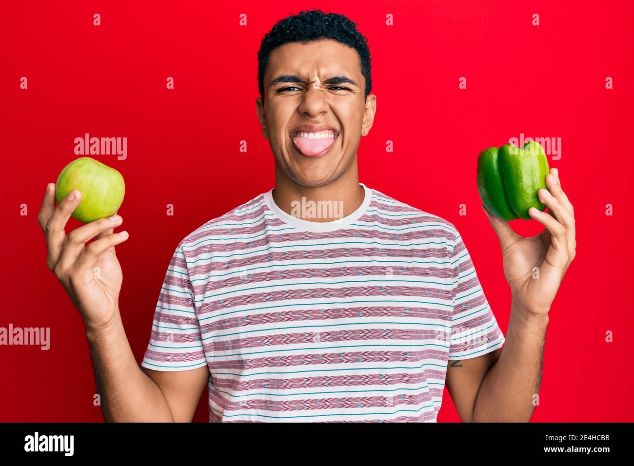 Young arab man holding green apple and pepper sticking tongue out happy with funny expression. Stock Photo