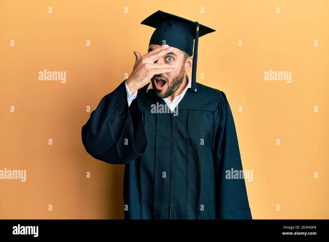 Young hispanic man wearing graduation cap and ceremony robe peeking in ...