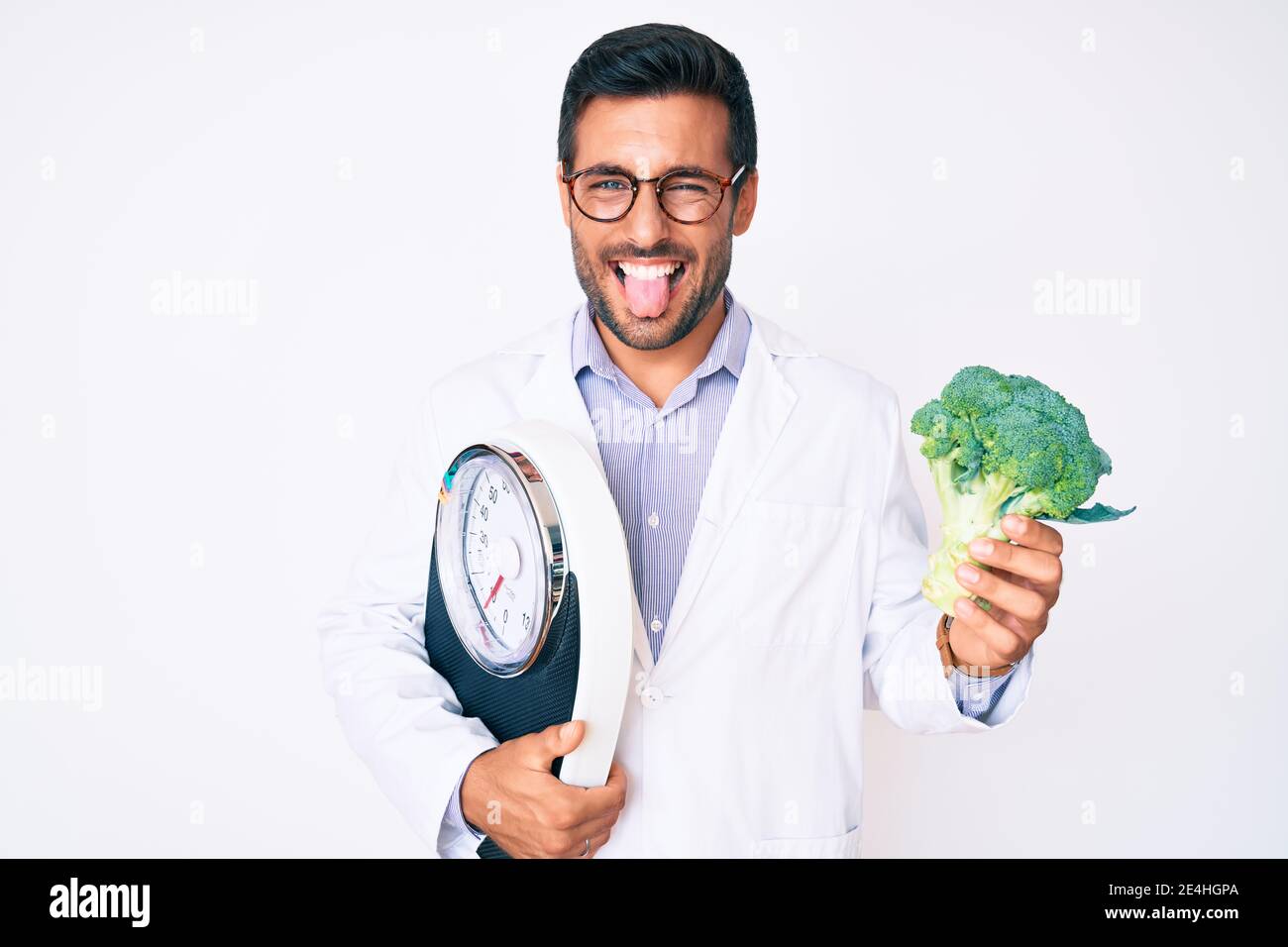 Young hispanic man as nutritionist doctor holding weighing machine and broccoli sticking tongue out happy with funny expression. Stock Photo