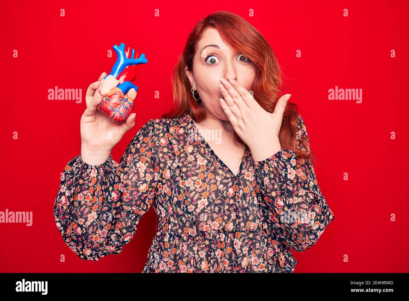 Young beautiful redhead woman asking for health care holding heart over read background covering mouth with hand, shocked and afraid for mistake. Surp Stock Photo