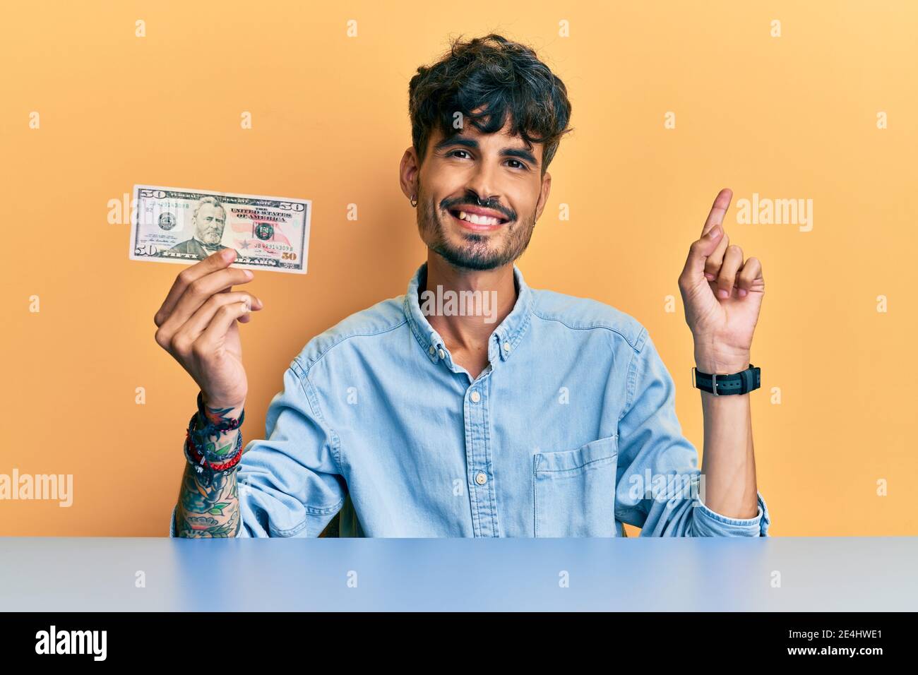 Young hispanic man holding 50 dollar banknote sitting on the table smiling happy pointing with hand and finger to the side Stock Photo