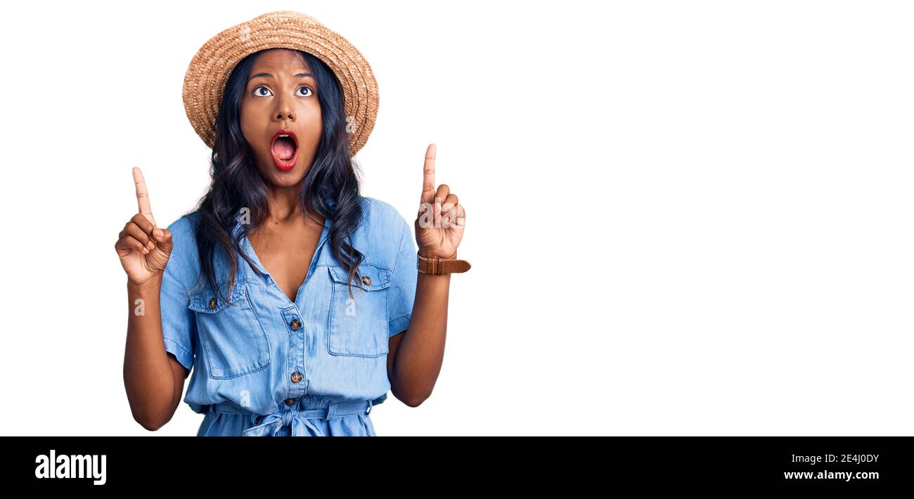 Young indian girl wearing summer hat amazed and surprised looking up and pointing with fingers and raised arms. Stock Photo