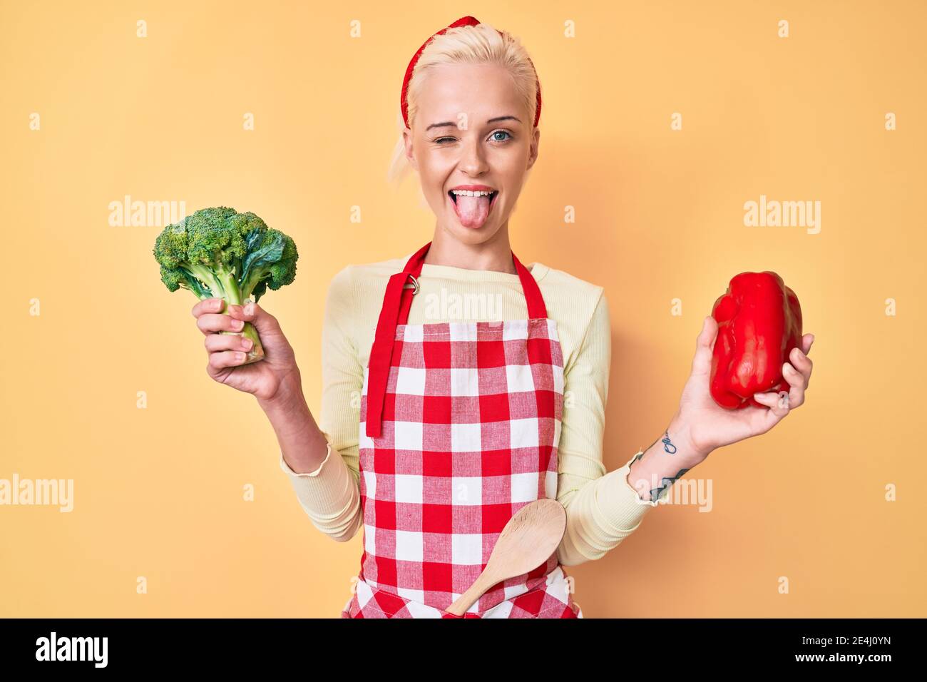 Young blonde woman with tattoo wearing cook apron holding broccoli and red pepper sticking tongue out happy with funny expression. Stock Photo
