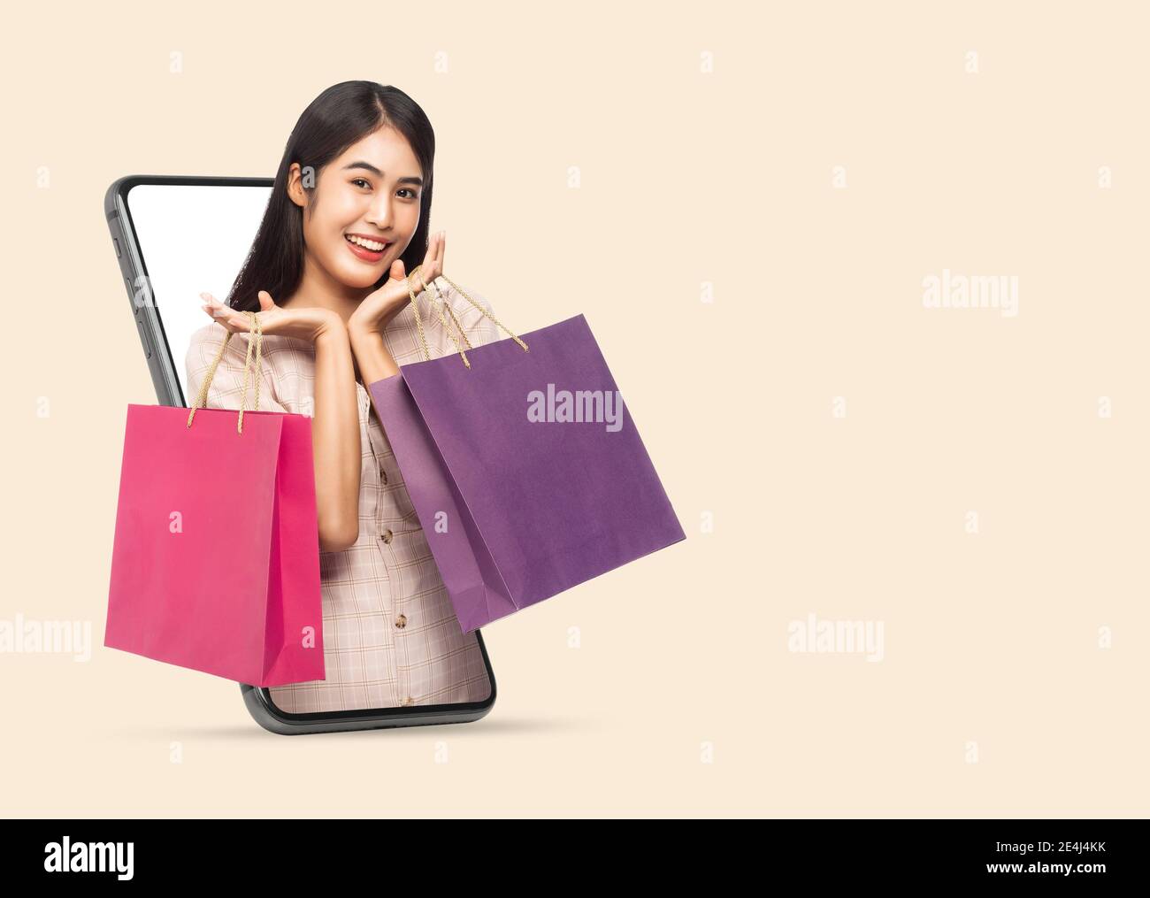 Young smiling beautiful asian woman making online shopping and holding shopping bags out through mobile phone isolated on beige background with clippi Stock Photo