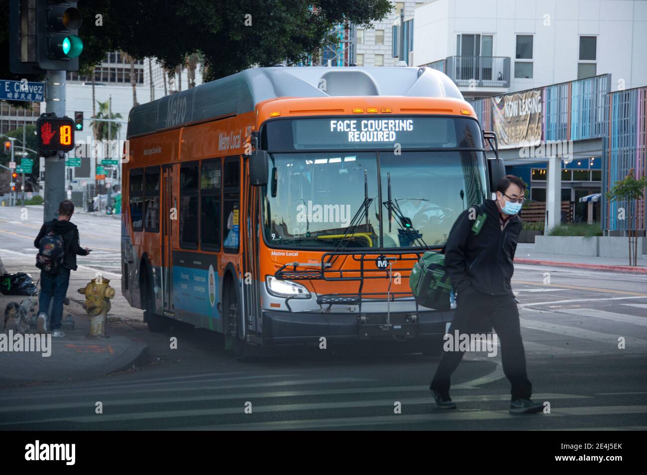 A young Asian man wearing a mask crosses the street, Behind him a bus states that masks must be worn in the bus. LA, CA, USA. Stock Photo