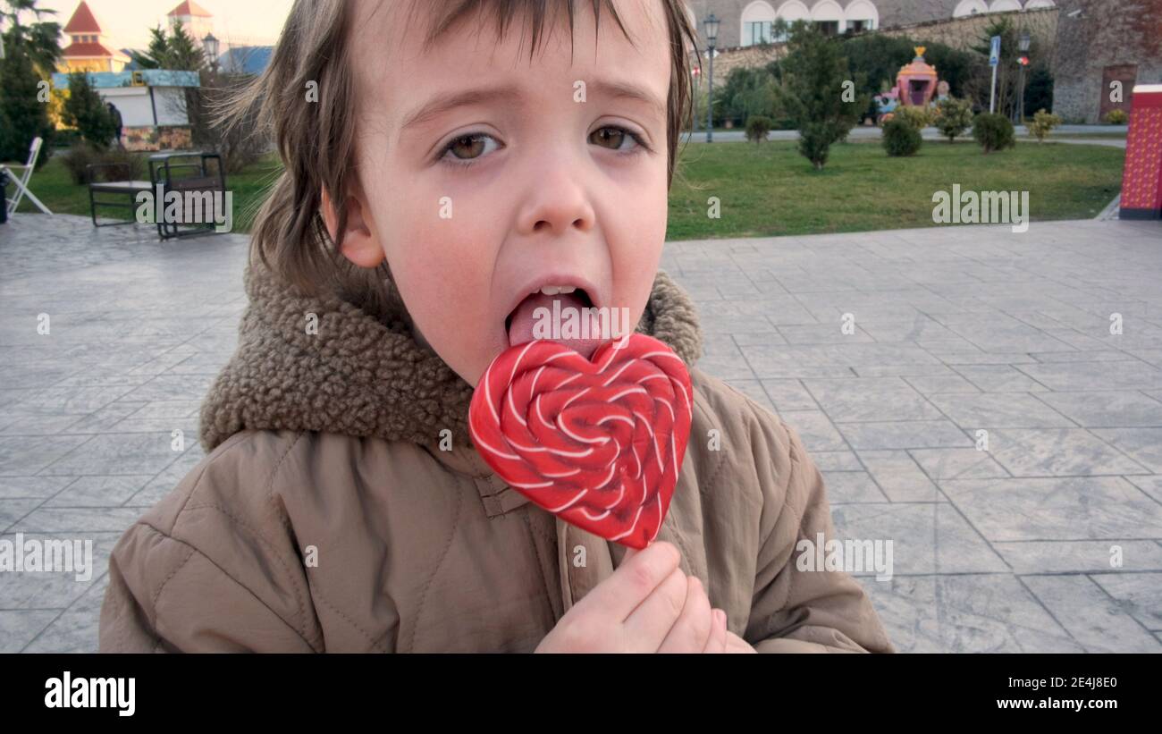 Funny little boy in warm brown jacket licks large heart shaped lollipop spending time in city park on autumn day closeup Stock Photo