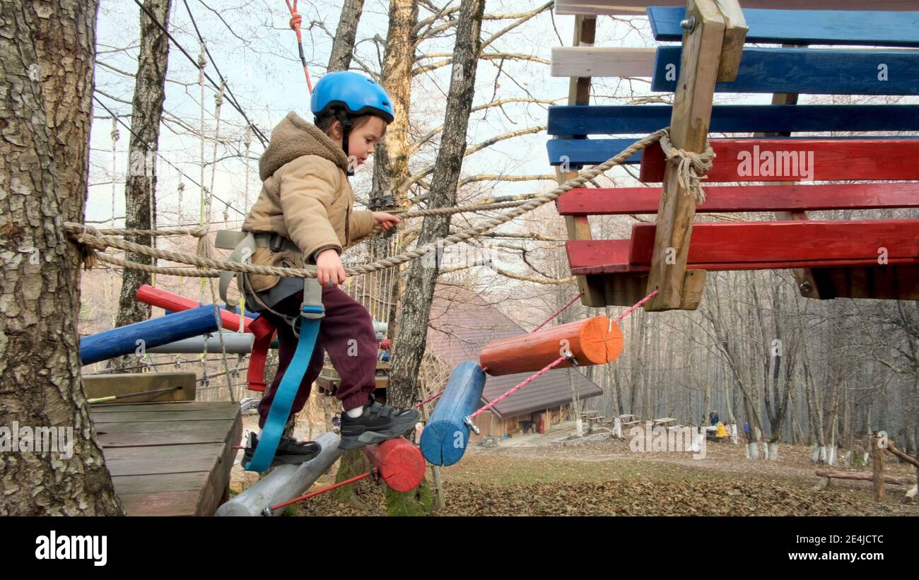 Brave toddler boy in protective helmet with safety belt goes up log stairs spending time at high rope course on autumn day Stock Photo