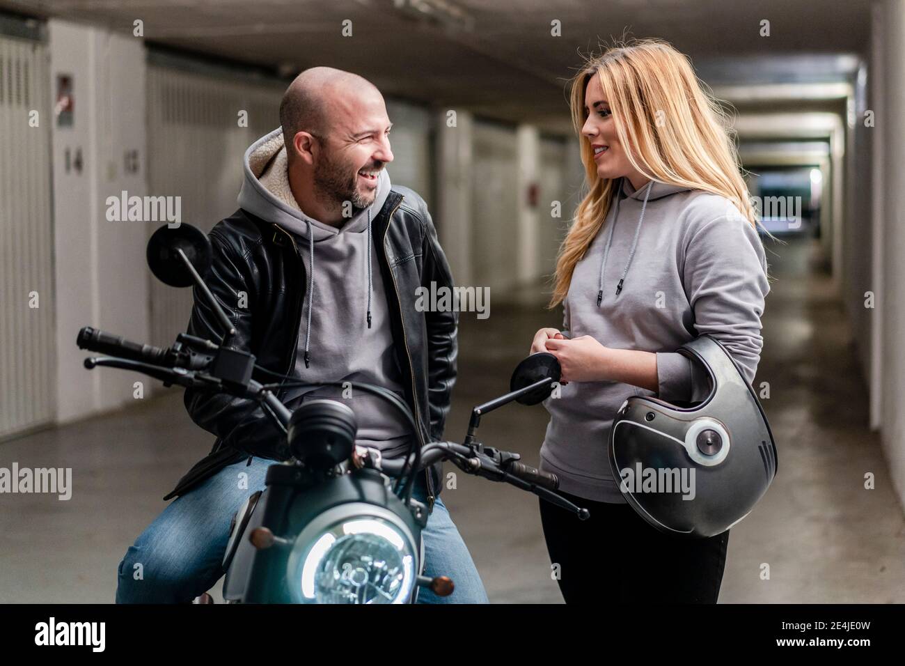 Smiling woman talking with boyfriend sitting on motorcycle in parking lot Stock Photo