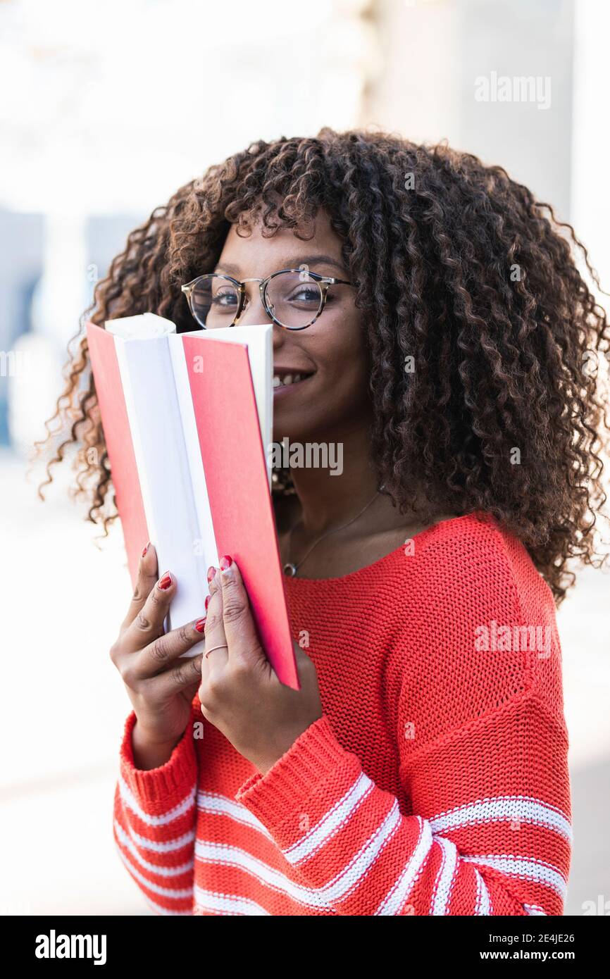 Happy young woman in warm clothes covering face with book Stock Photo