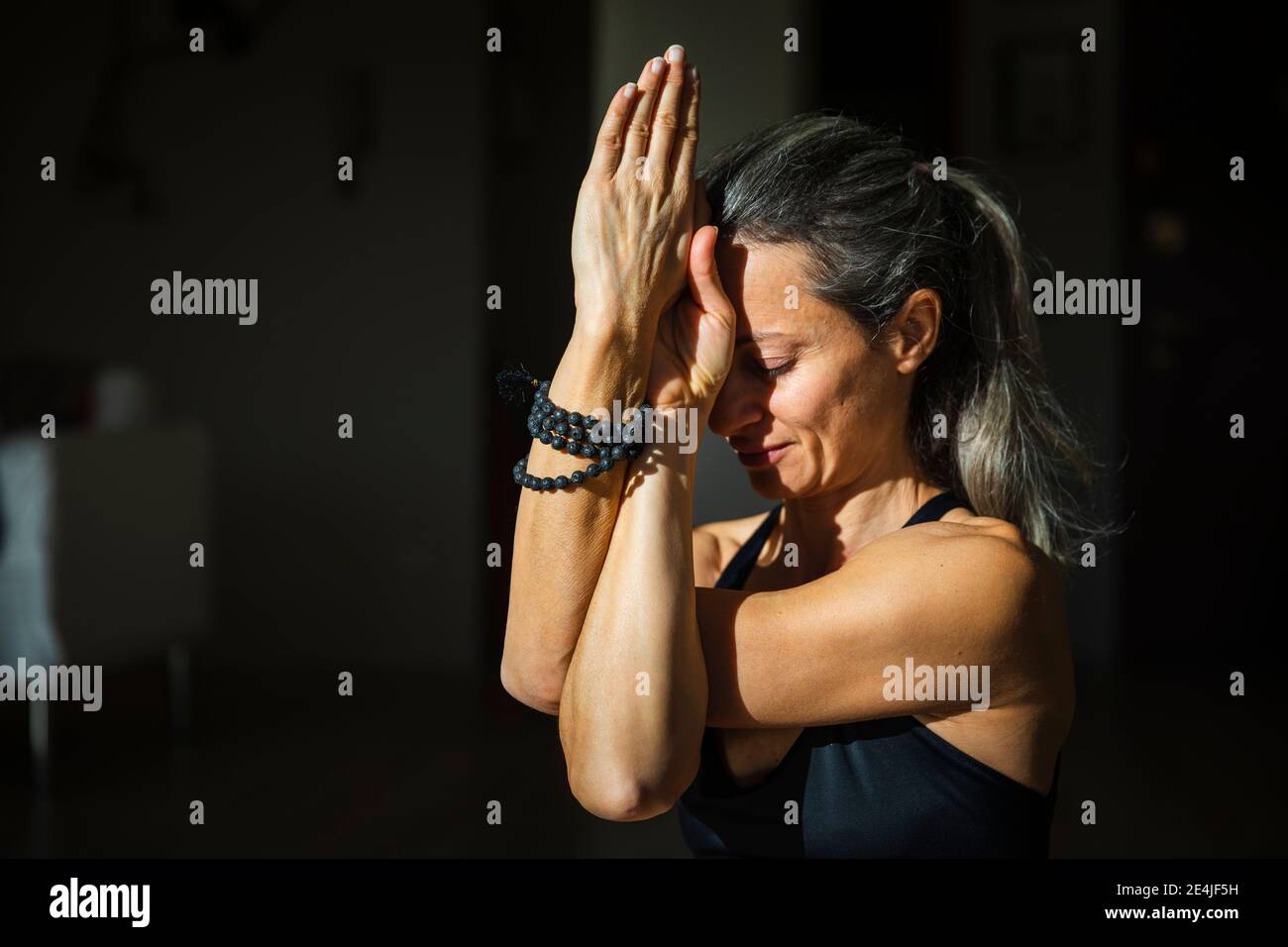 Mature woman practicing yoga with twisting arms at home on sunny day Stock Photo