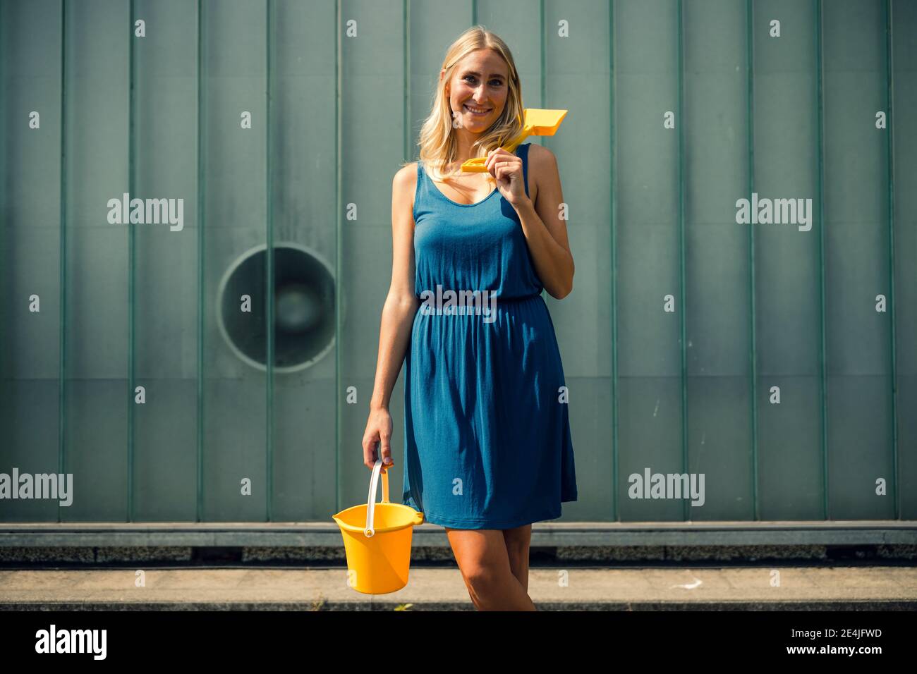 Smiling woman with sand bucket and shovel standing on footpath against wall Stock Photo