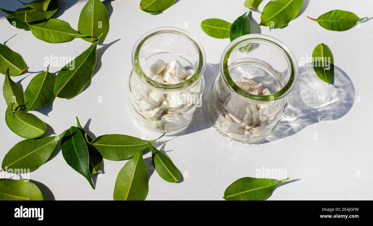 Two glass jars with mineral supplements pills. Green leaves. White background. Transparent glass shadow. Health topics. Stock Photo