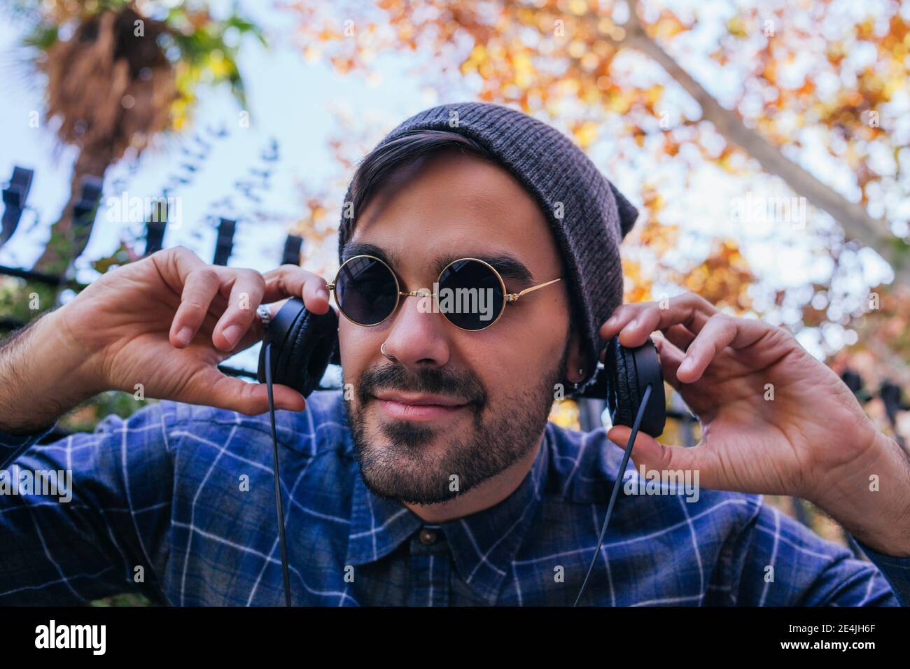 Smiling fashionable man wearing nose ring holding headphones in public park Stock Photo