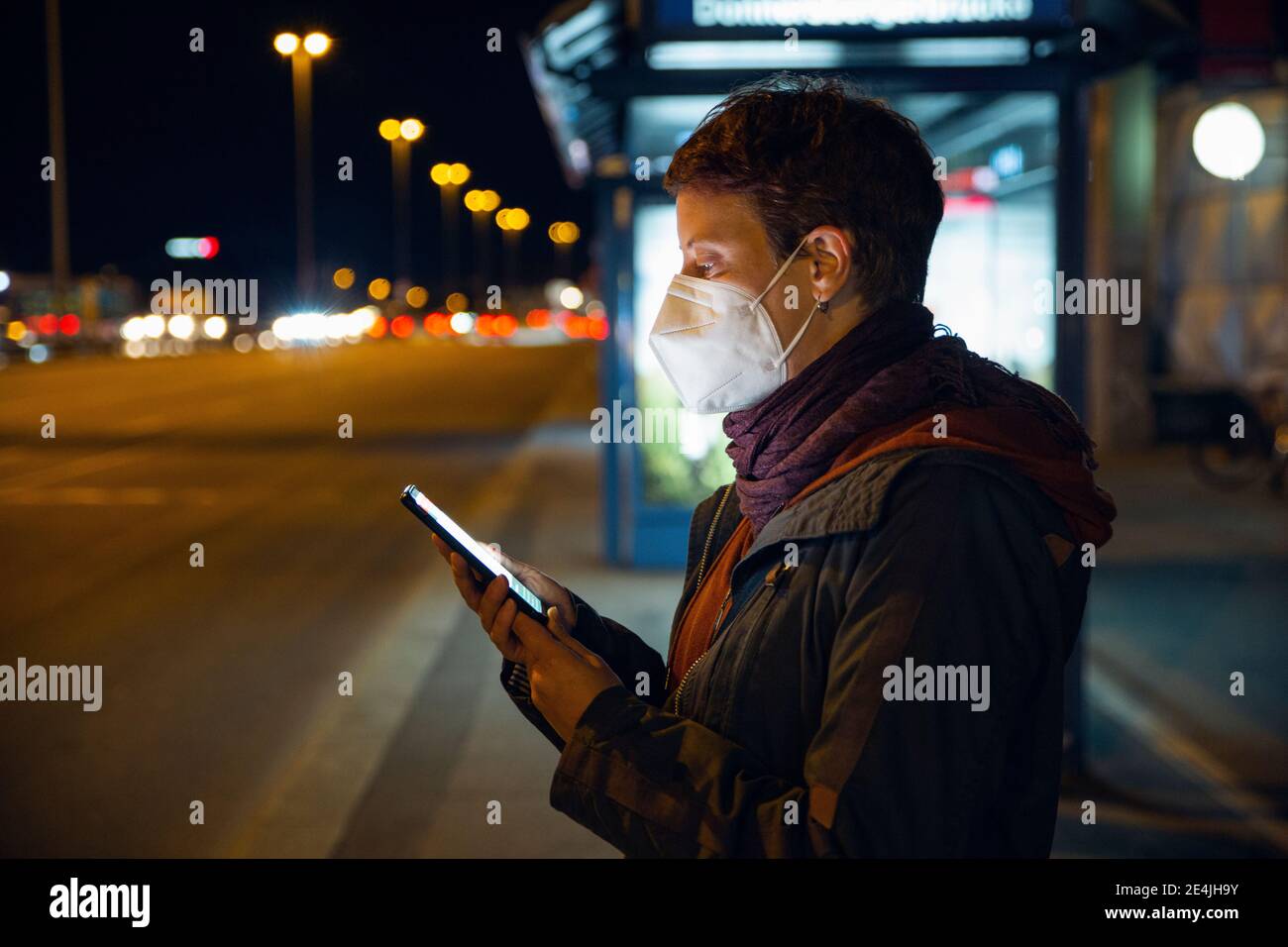 Woman wearing FFP2 mask using smart phone while waiting for bus in city Stock Photo