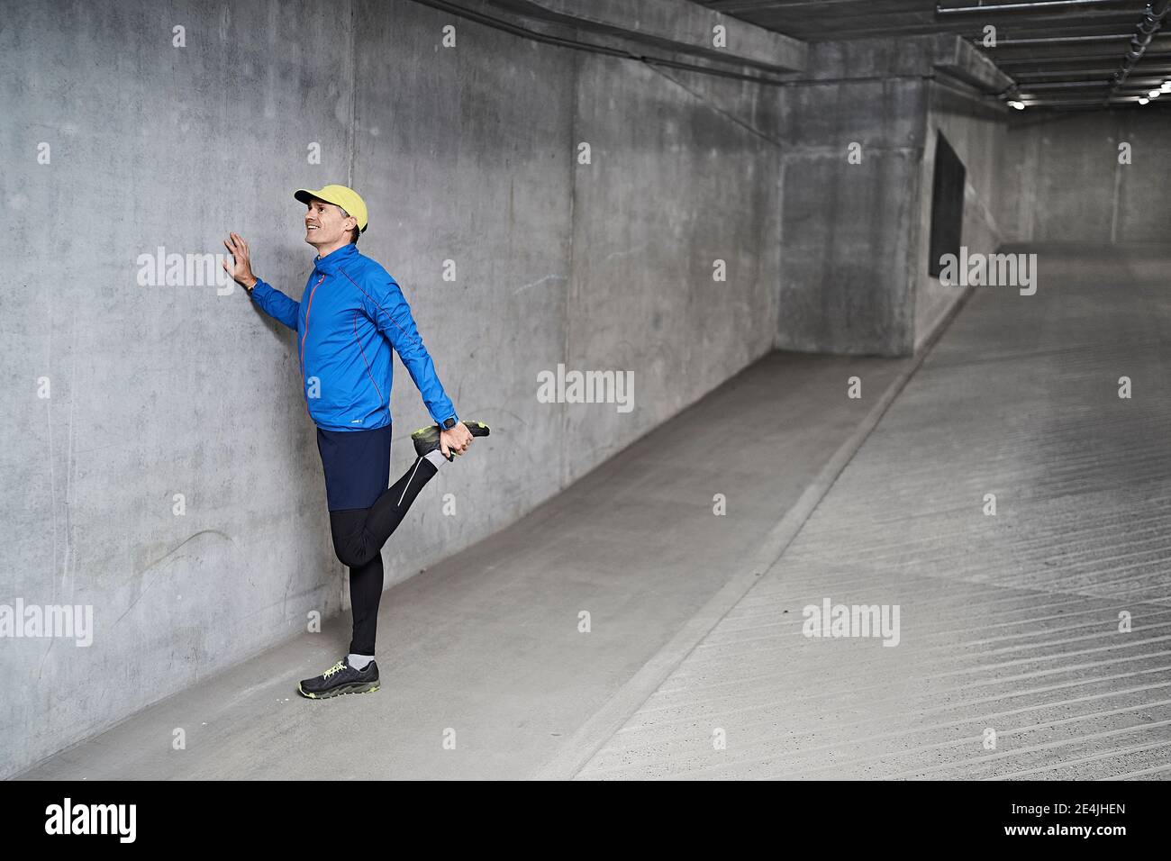 Smiling mature man stretching leg by gray wall in parking garage Stock Photo