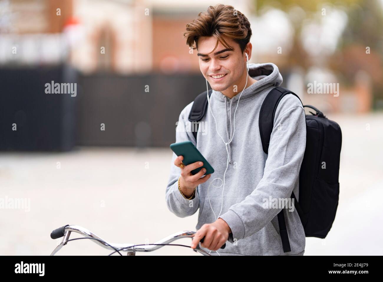 Smiling fashionable young man using smart phone while wheeling bicycle in city Stock Photo