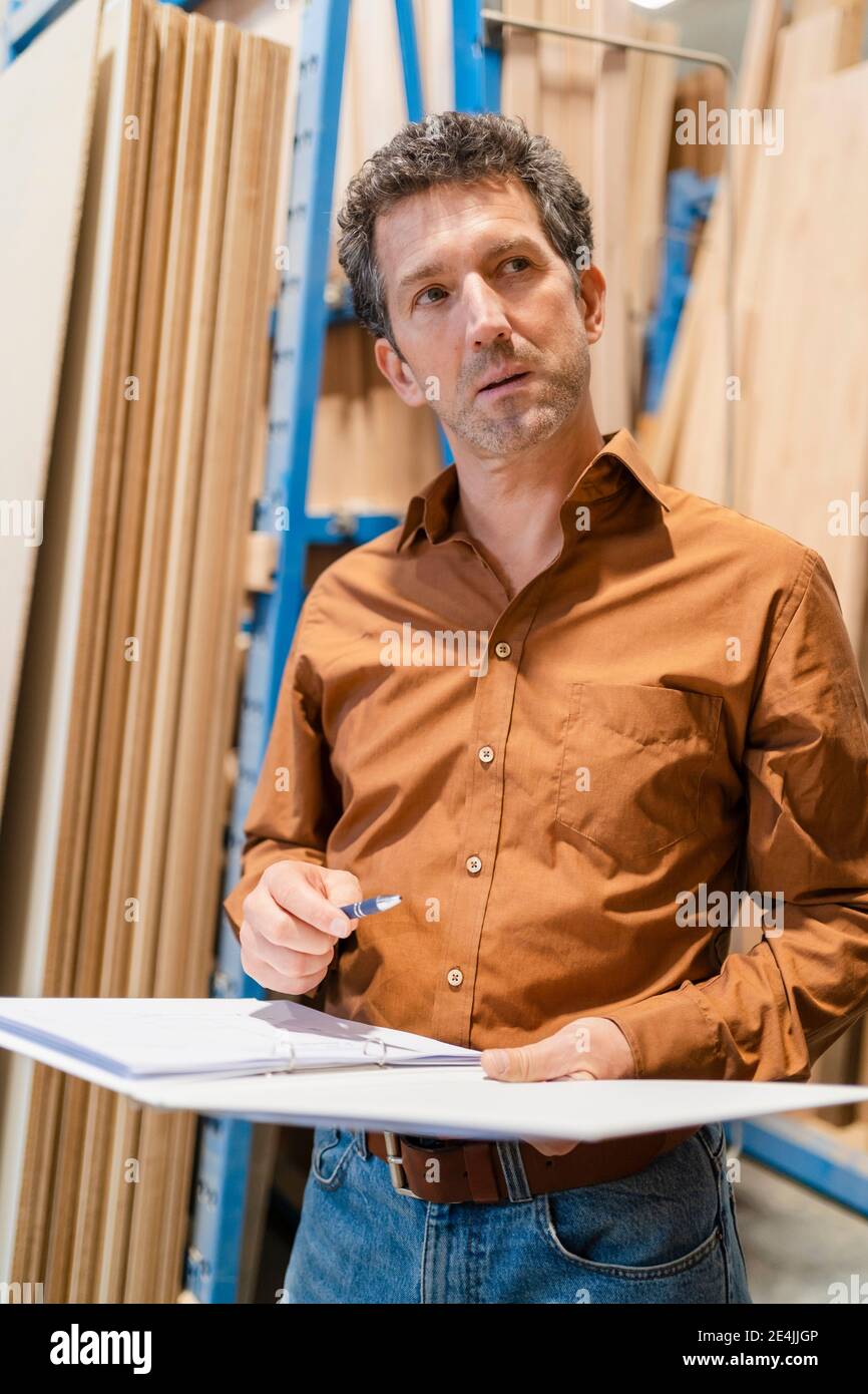 Portrait of carpenter standing with ring binder in front of shelves with wooden planks Stock Photo