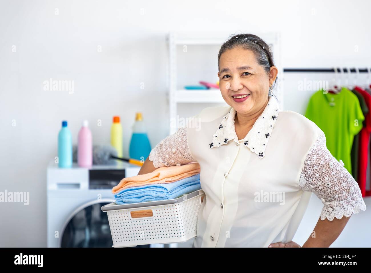 Happy smiling Asian senior elderly female woman carrying clothe basket doing laundry with washing machine, house cleaning and housekeeping, looking at Stock Photo