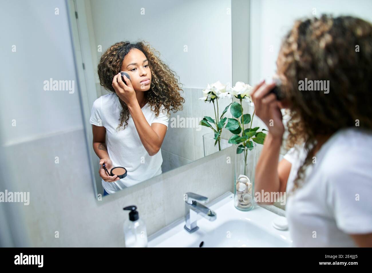 Young woman applying face powder with make-up brush while looking at mirror reflection in bathroom Stock Photo