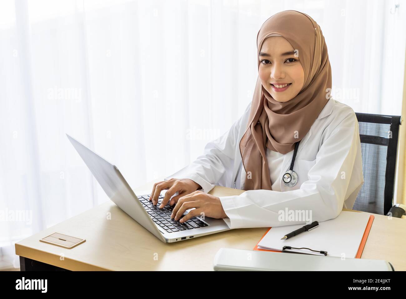 Confident Muslim female woman medical doctor sitting at desk, working typing with laptop computer in clinic hospital office; looking at camera Stock Photo