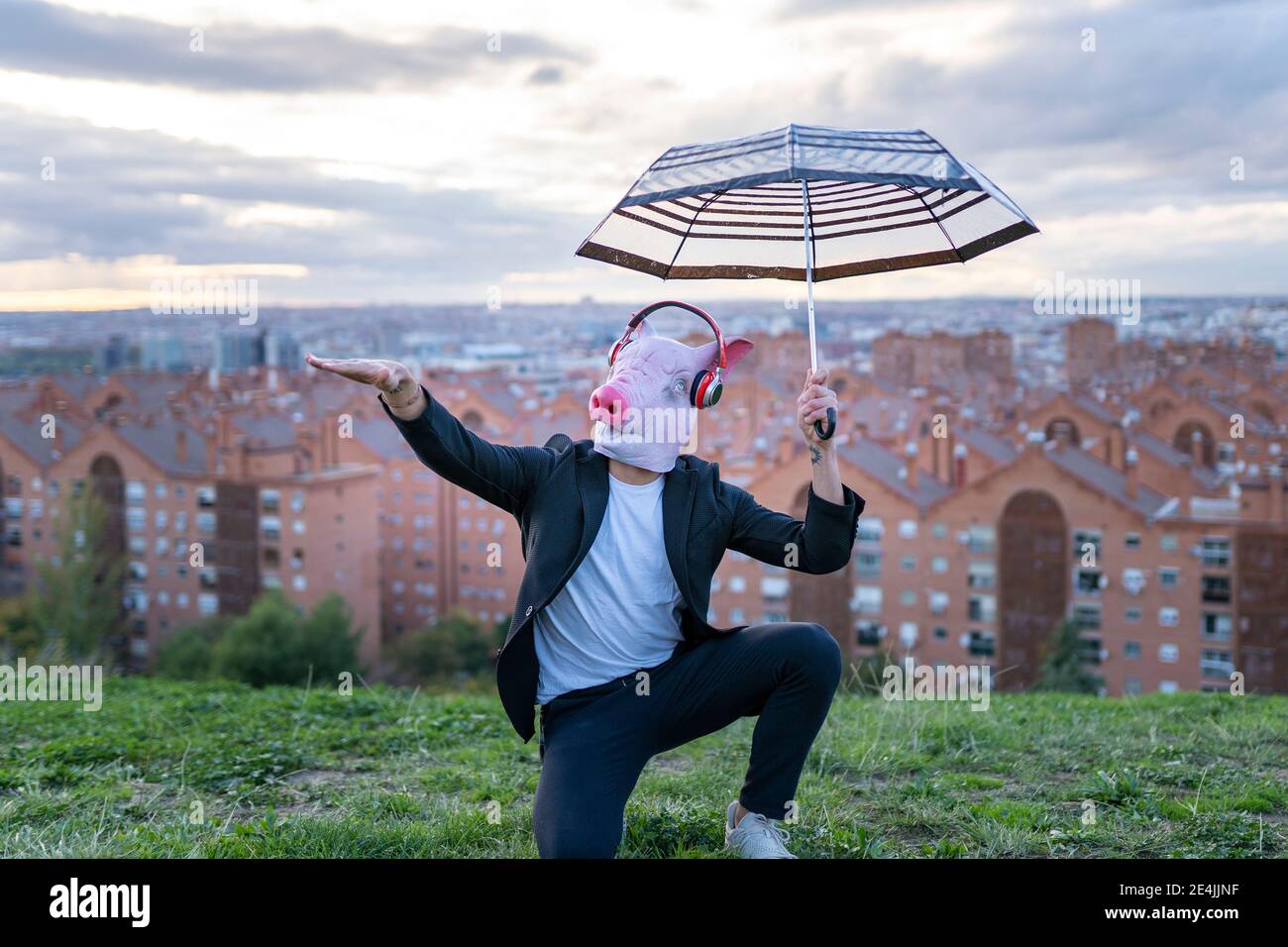 Male professional wearing pig mask holding umbrella gesturing while kneeling against sky at sunset Stock Photo