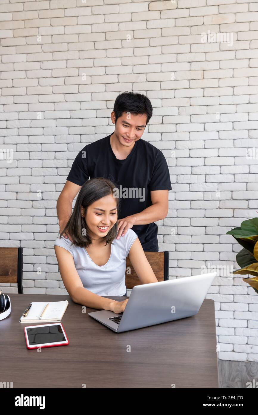 Asian man husband motivate support wife woman freelancer working at home, sitting at desk dining table in living room Stock Photo