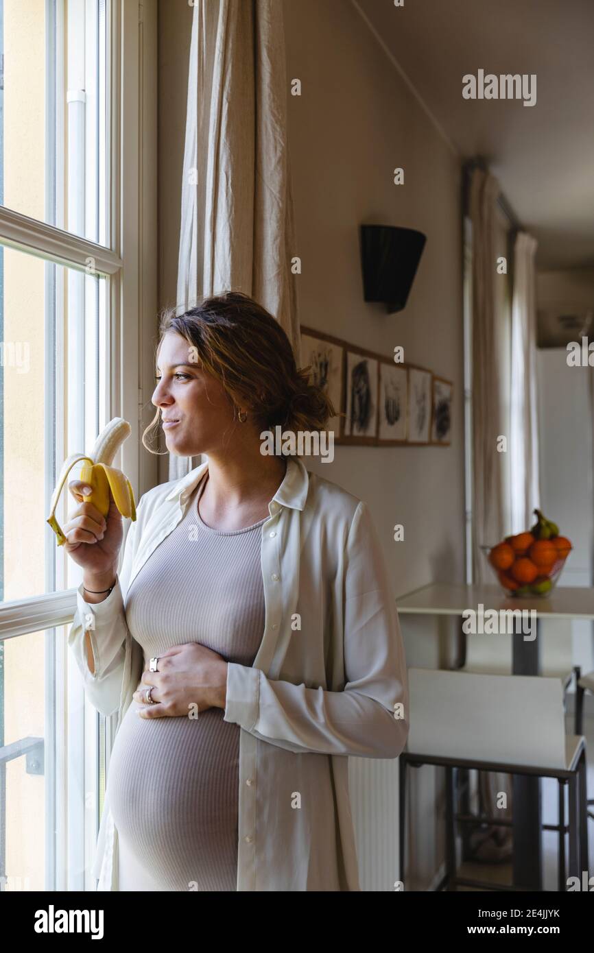 Young pregnant woman eating banana while looking through window at home
