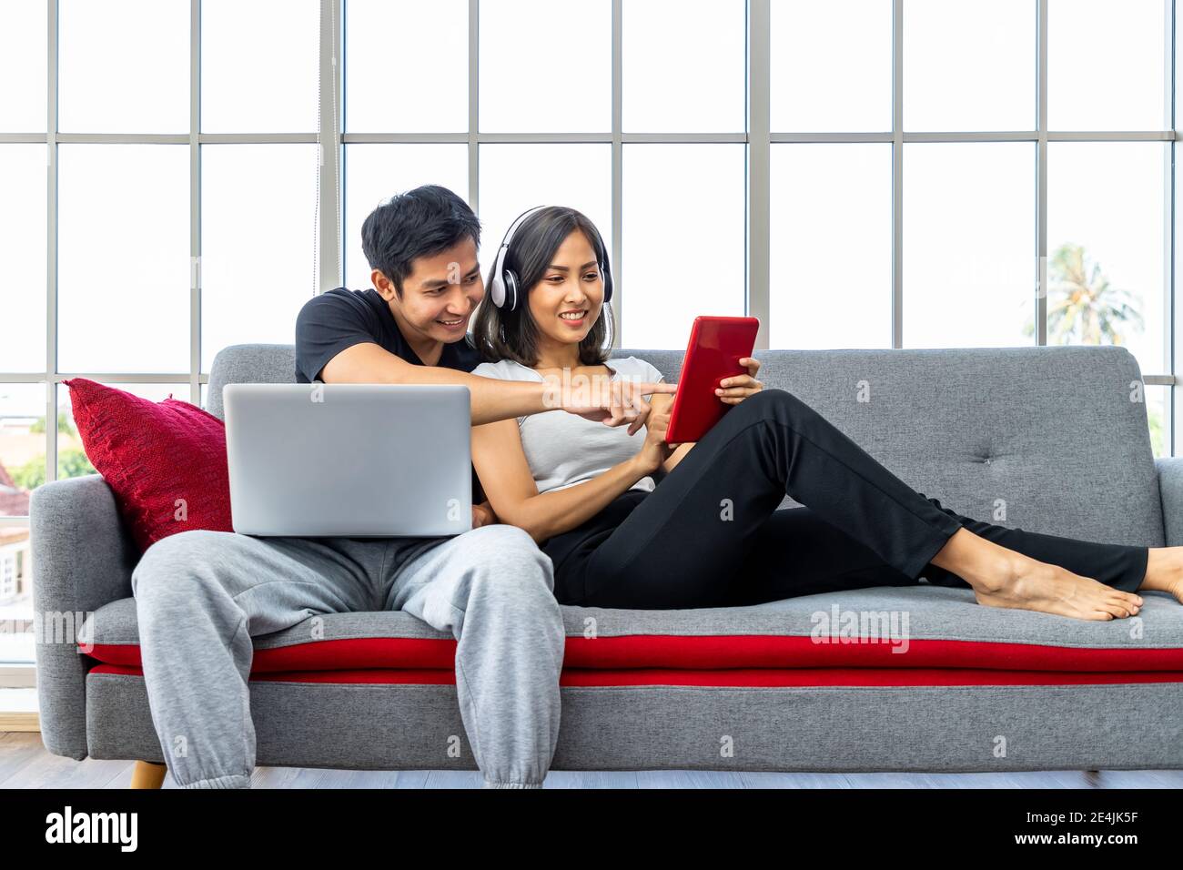 Young Asian couple relaxing sitting on sofa using laptop computer and digital tablet working from home Stock Photo
