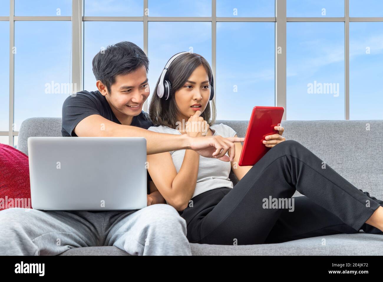 Young Asian couple relaxing sitting on sofa using laptop computer and digital tablet working from home Stock Photo
