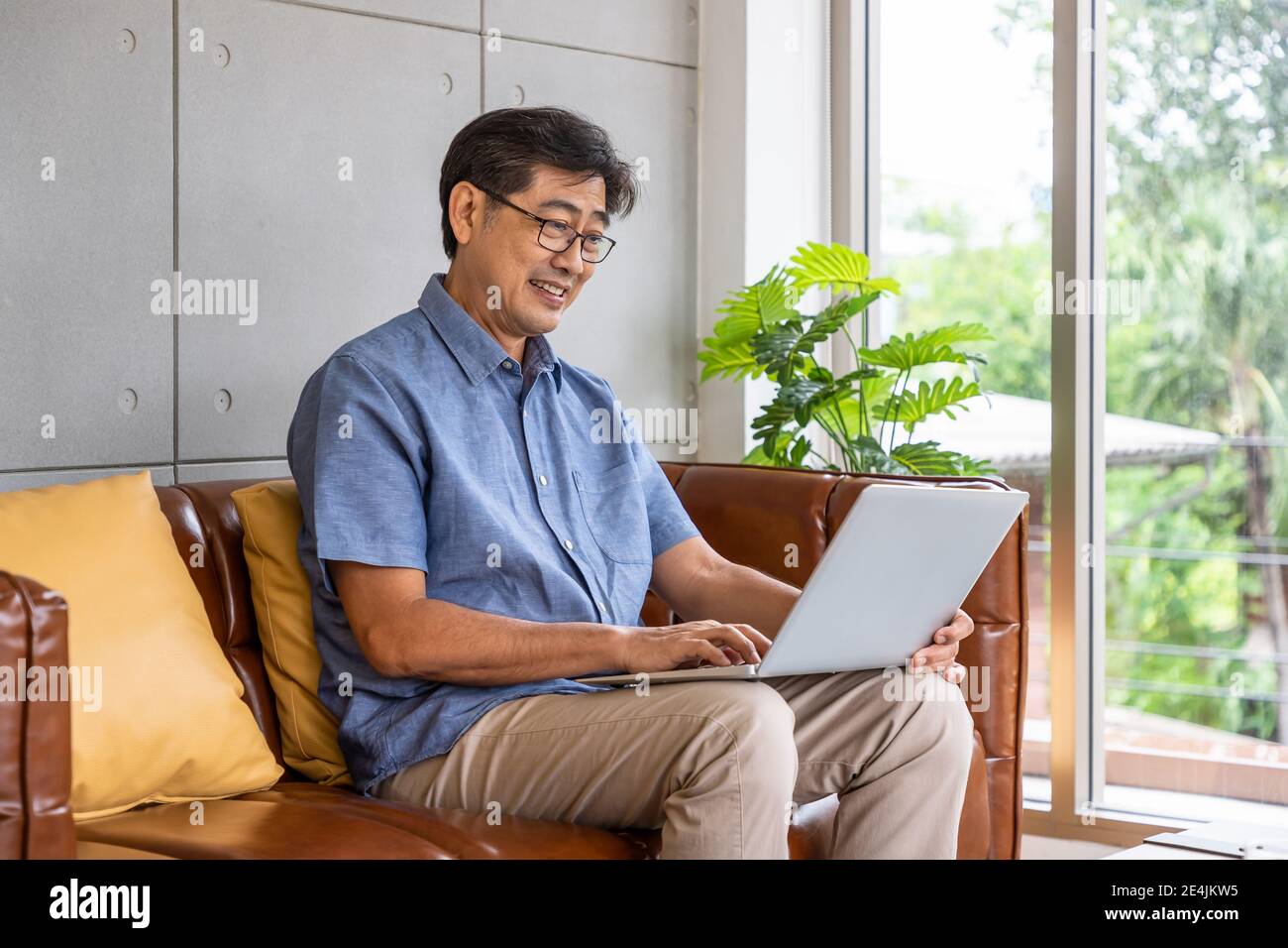 Happy Asian senior retired man sitting on couch sofa, using laptop computer Stock Photo