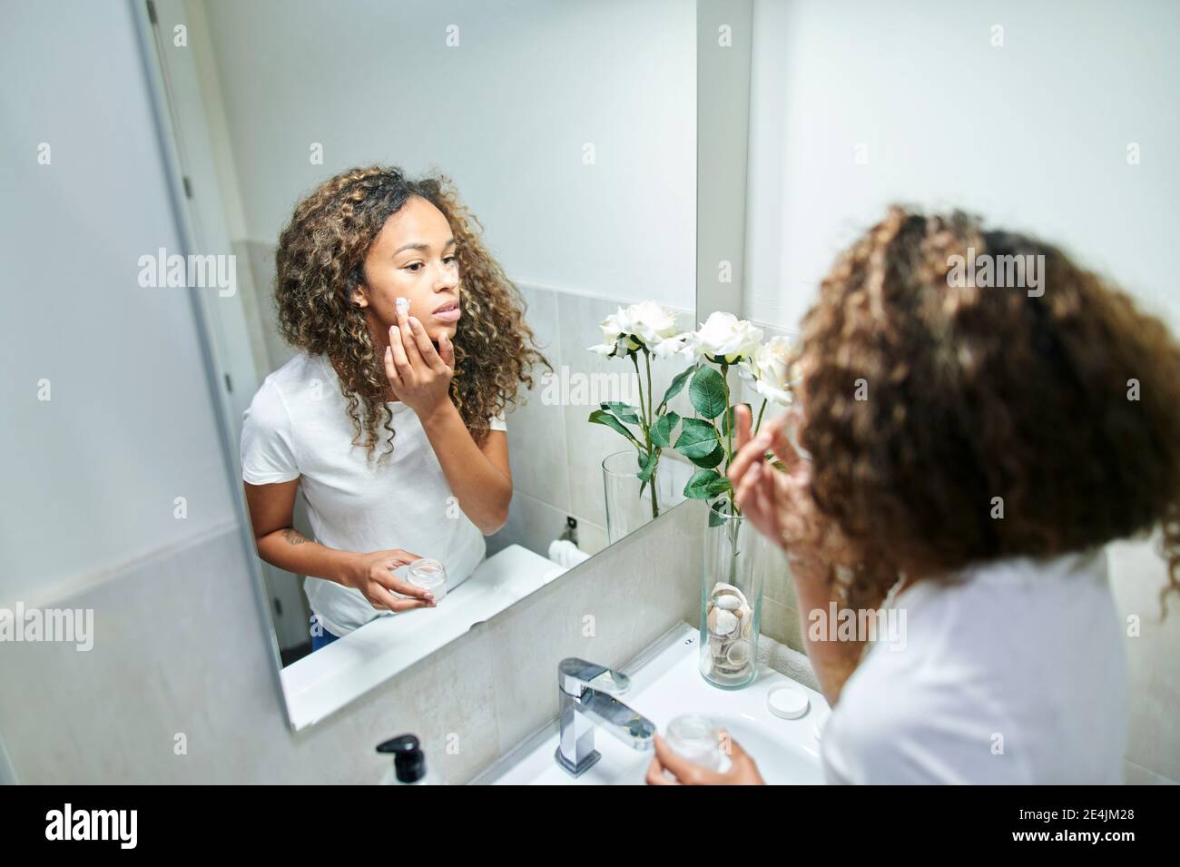 Beautiful woman applying facial cream while looking at mirror reflection in bathroom Stock Photo