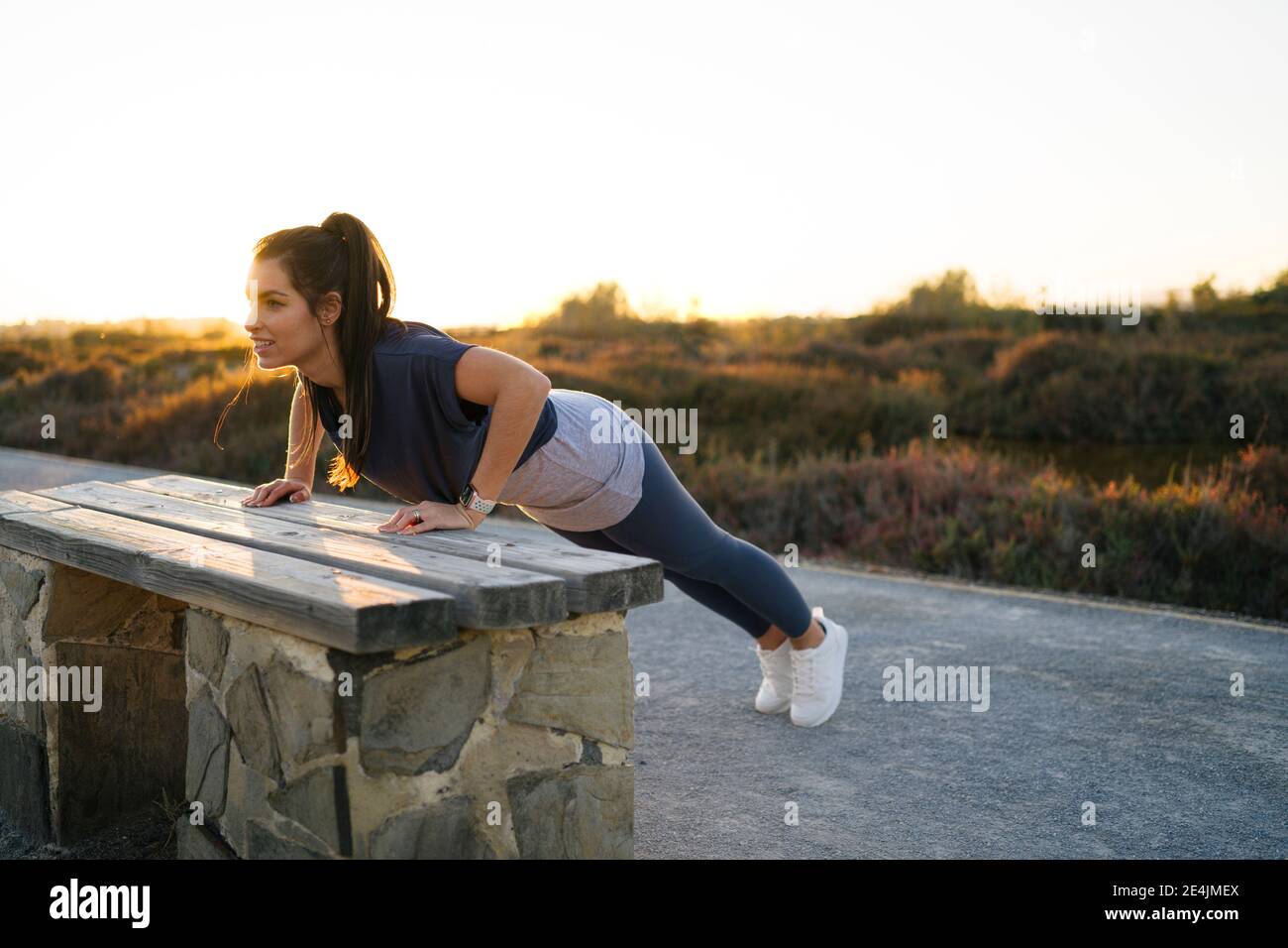 Young woman practicing push-ups on bench against clear sky at sunset Stock Photo