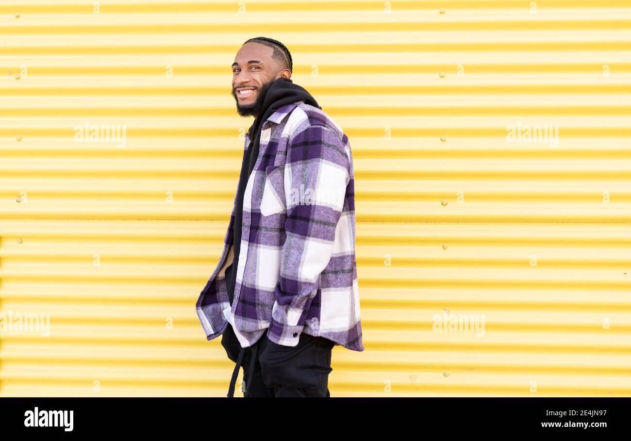 Smiling young man with hands in pockets against yellow corrugated iron Stock Photo