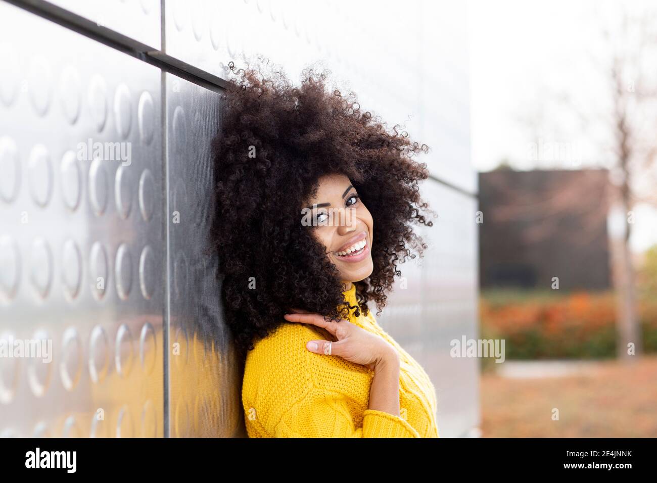 Curly hair woman smiling while leaning on silver wall Stock Photo