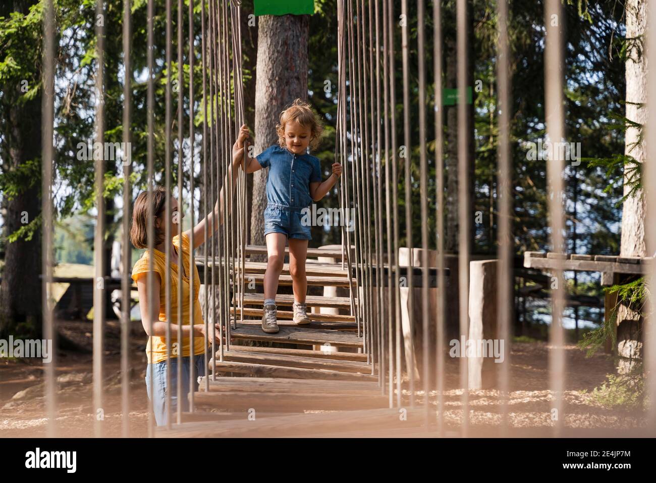 Mother assisting little daughter walking across small suspension bridge on forest obstacle course Stock Photo