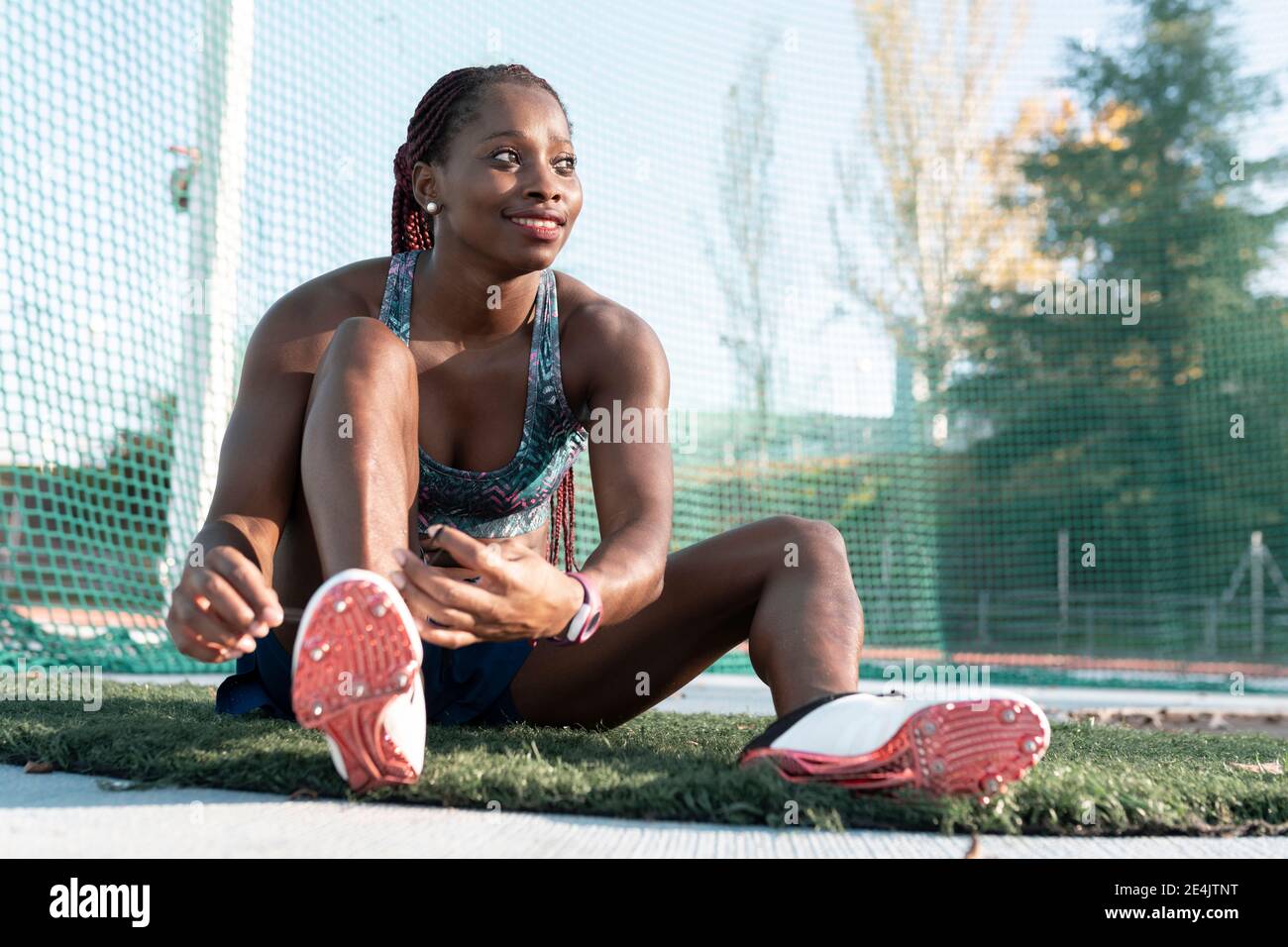 Smiling young sportswoman looking away while tying shoelace against net Stock Photo