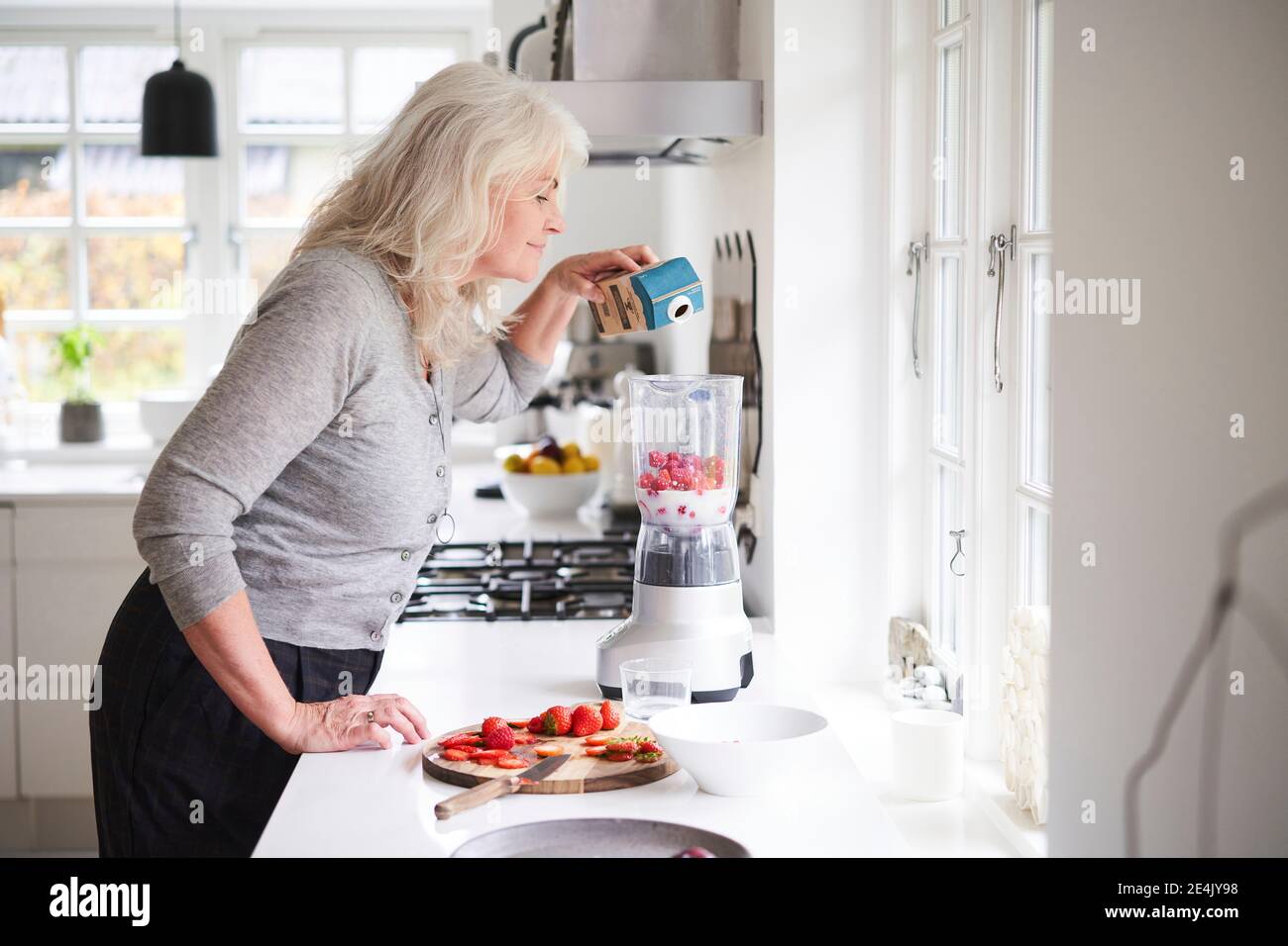 Senior woman pouring milk in processor while preparing strawberry smoothie at kitchen Stock Photo