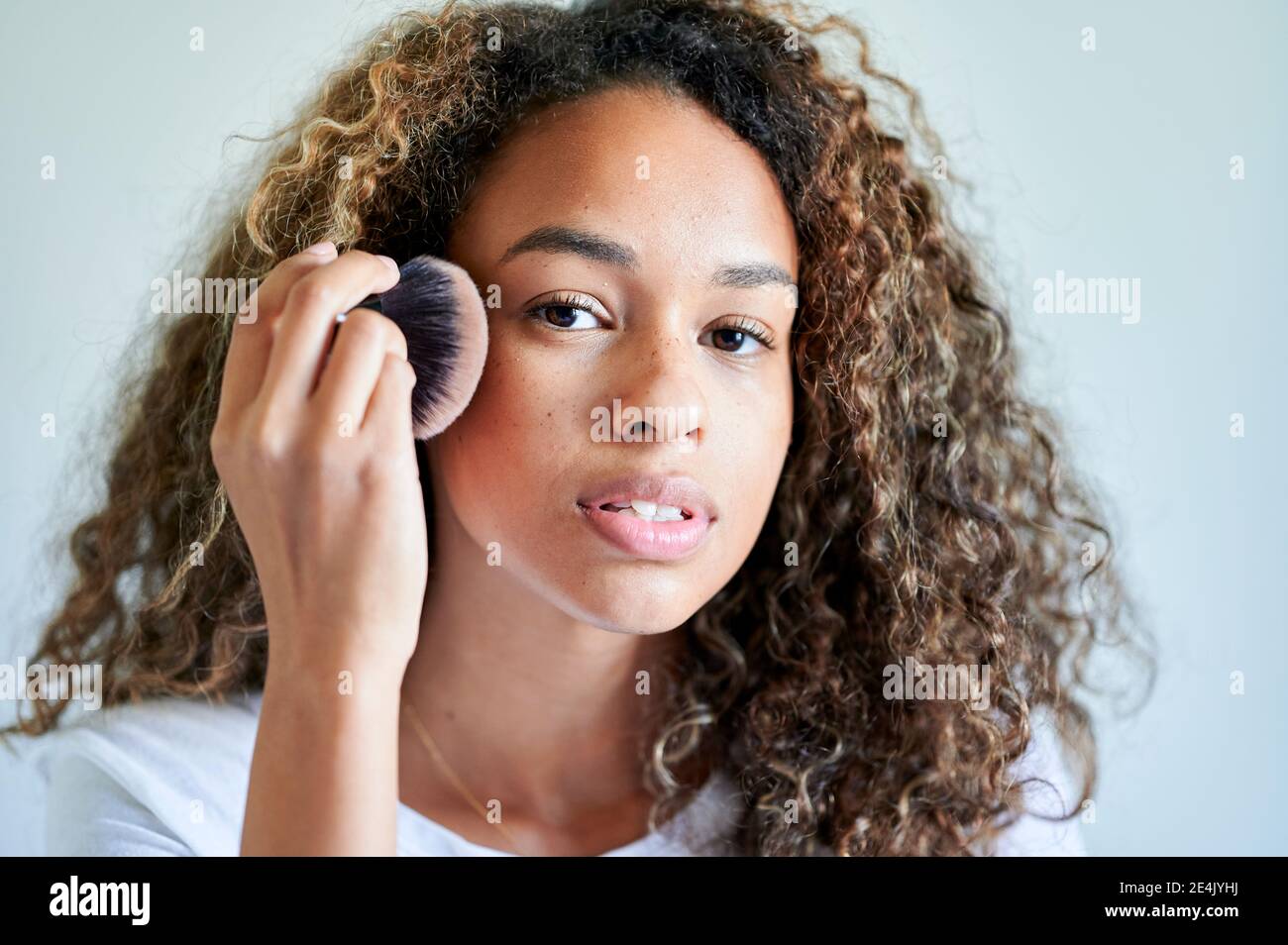 Young woman applying face powder with make-up brush against wall Stock Photo