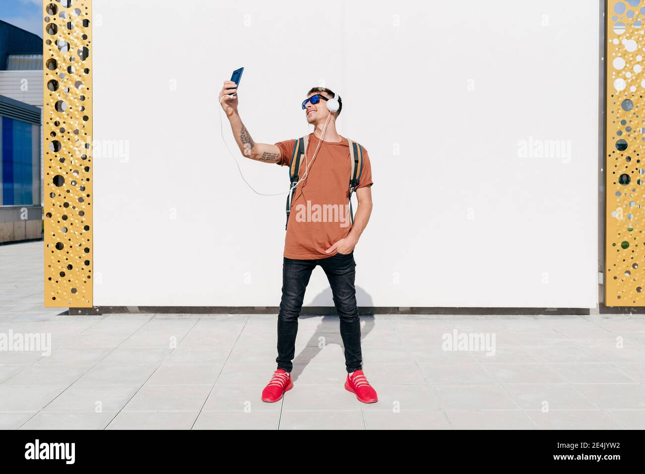 Smiling man wearing headphones taking selfie through mobile phone while standing with hands in pockets against wall Stock Photo