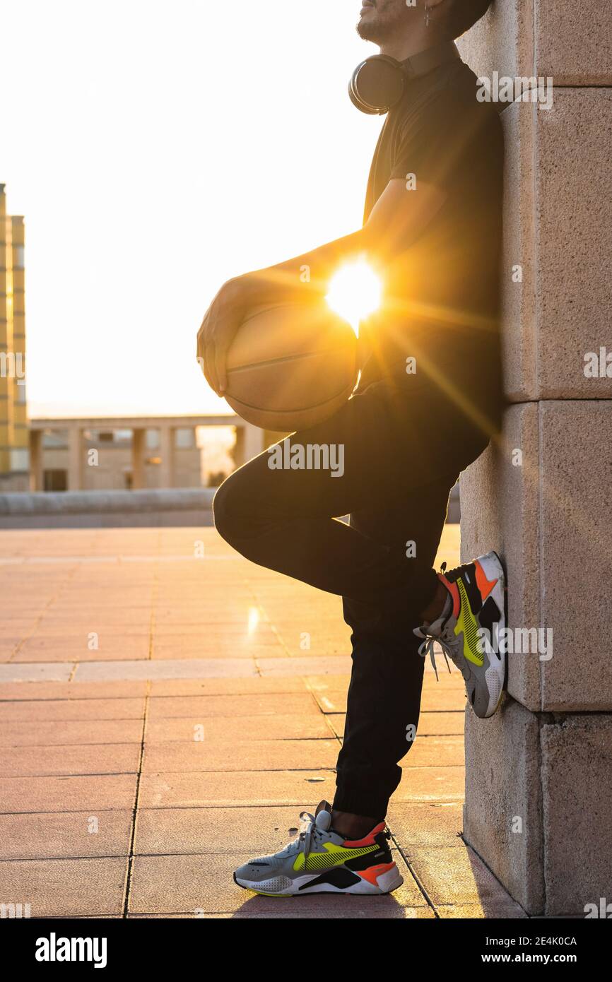 Sun shining through young man holding basketball while standing by wall at sunset Stock Photo